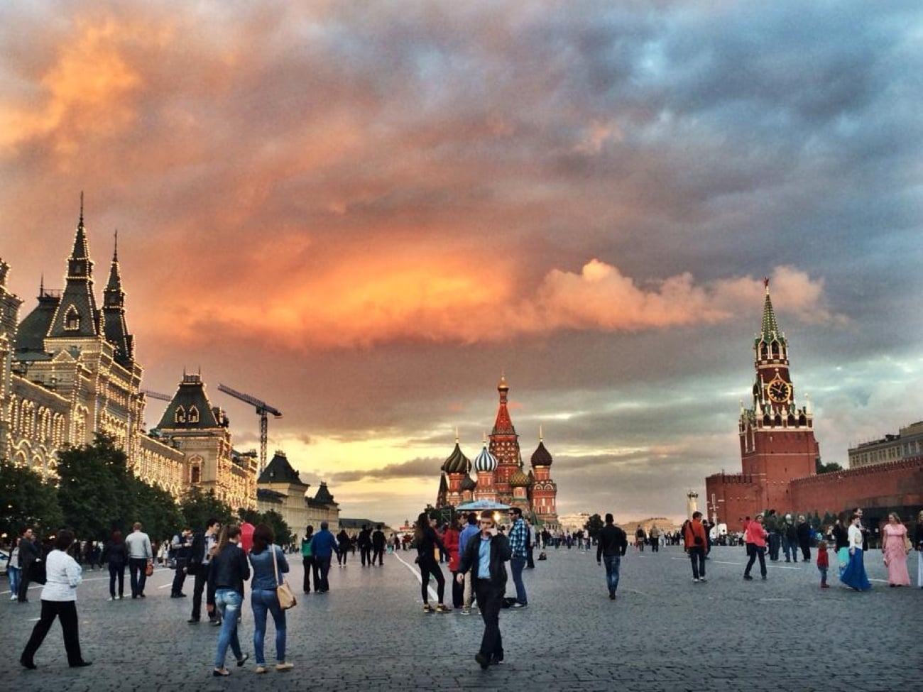 People walk across Red Square in Moscow at sunset, with St. Basil’s Cathedral and the Kremlin towers visible under a dramatic, colorful sky. The buildings are illuminated, and the plaza is busy with visitors exploring one of the Biggest Cities In The World.
