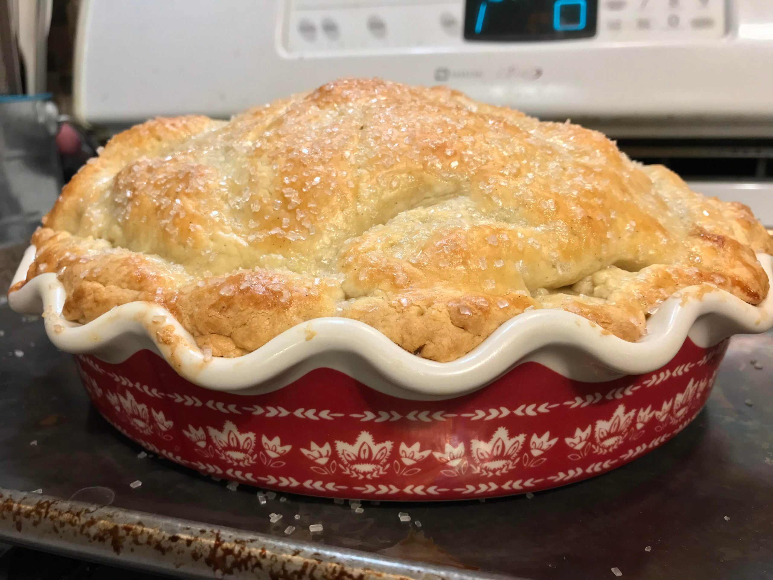 Golden-brown pie with a sugar-crusted top in a red and white decorative pie dish, sitting on a baking tray in front of an oven.