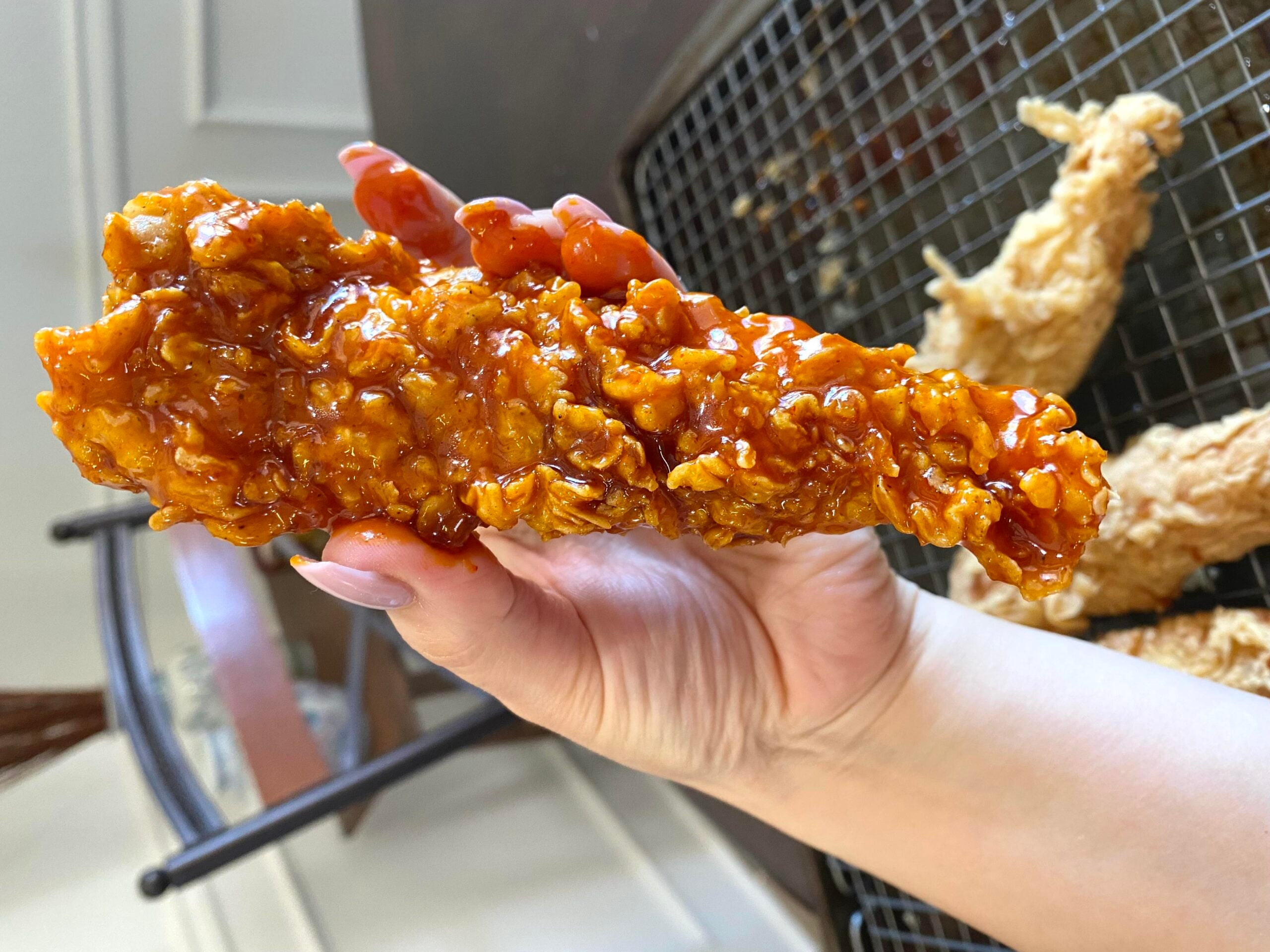 A hand holds a crispy fried chicken tender coated in a shiny, orange sauce. In the background, more tenders rest on a wire rack.