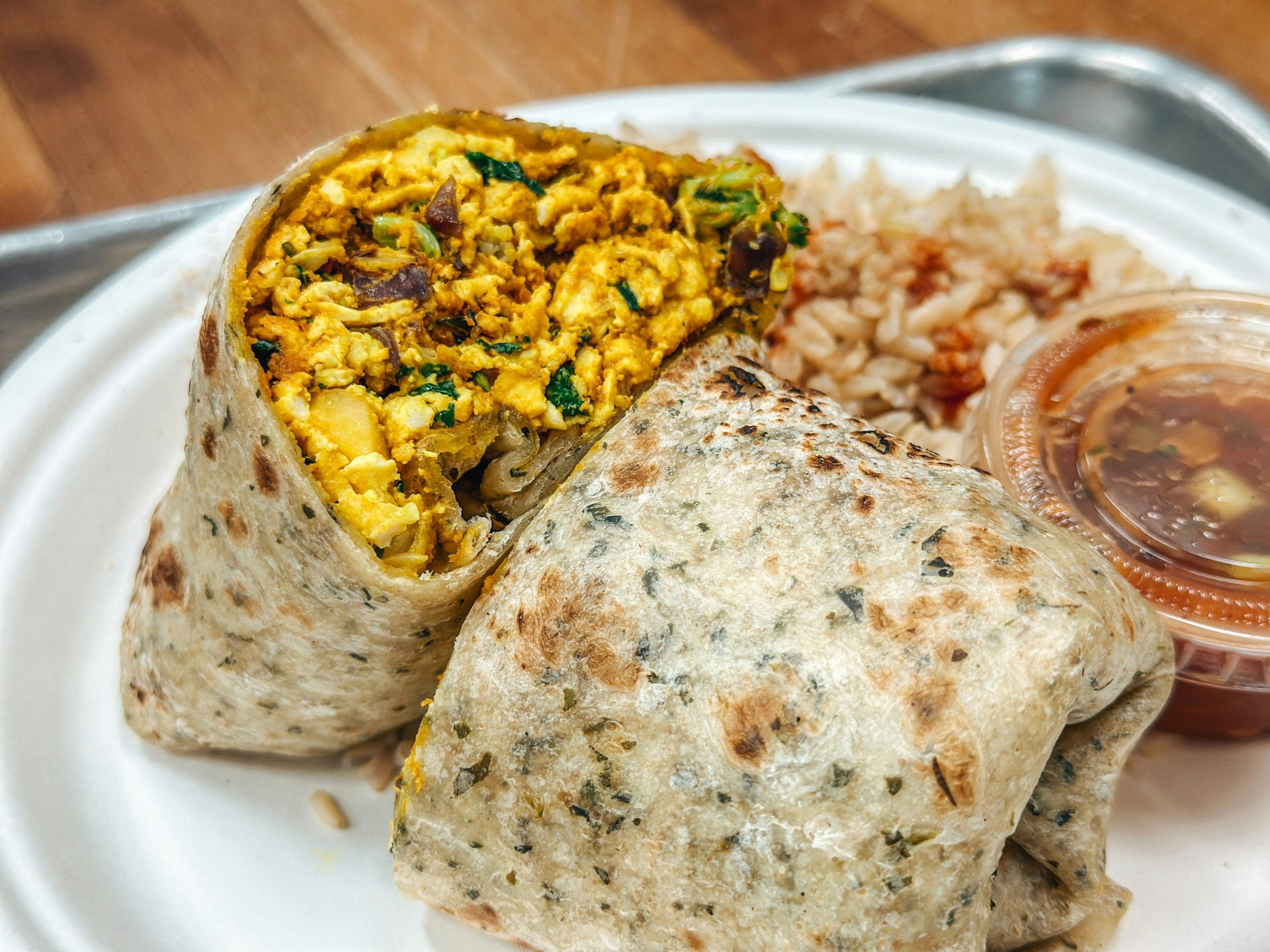 A close-up of a cut breakfast burrito filled with scrambled tofu and veggies, served with brown rice and a small container of salsa on a white plate.