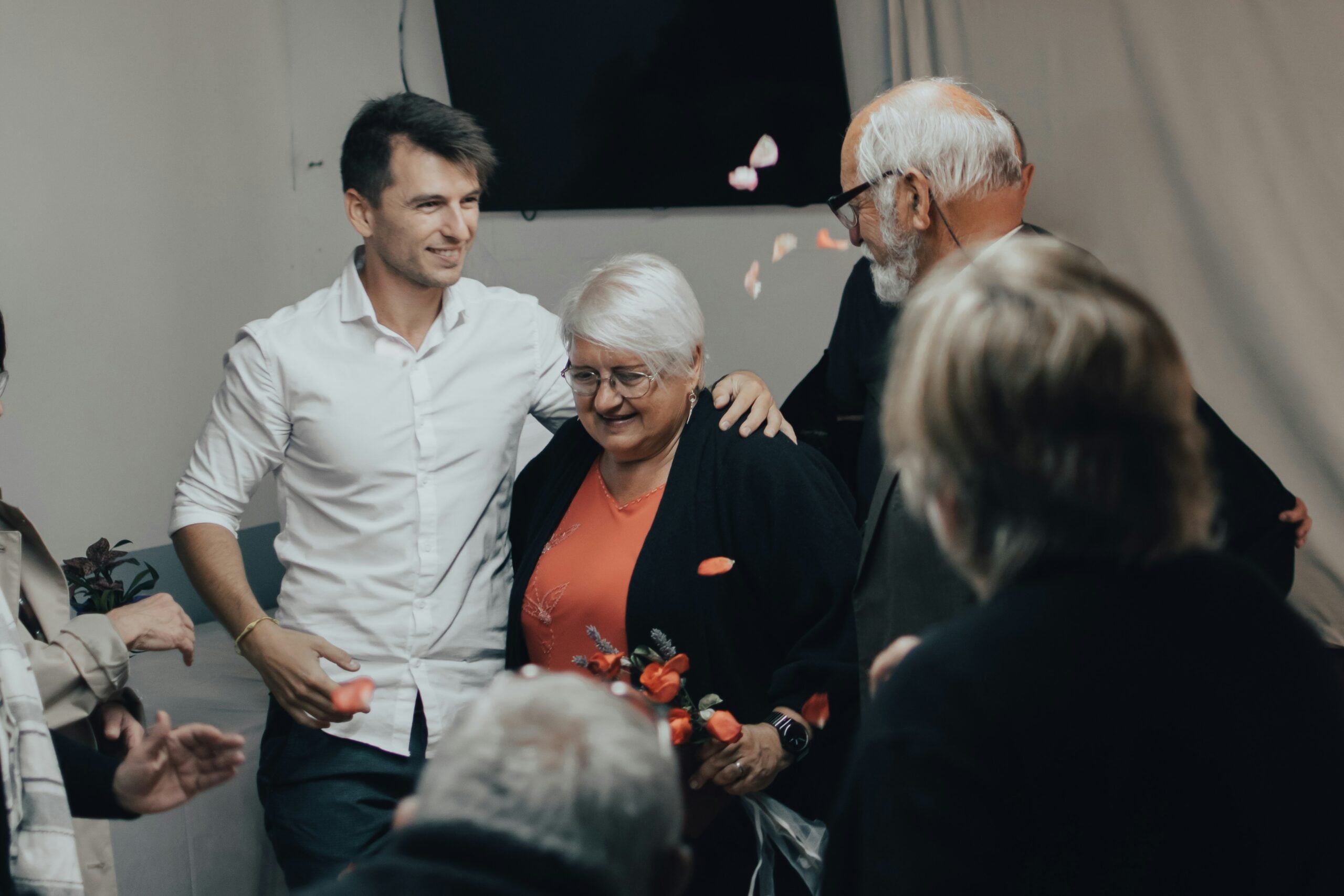 A group of people, including an older woman holding flowers and a younger man, stand together smiling as flower petals fall around them in a warmly lit indoor setting, celebrating her years of dedication and the retirement savings that will support her next chapter.
