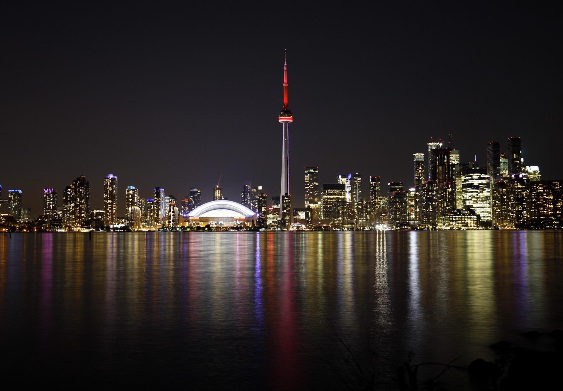 Toronto skyline at night with the CN Tower lit up in red, surrounded by illuminated buildings, and colorful reflections shimmering on the water in the foreground.
