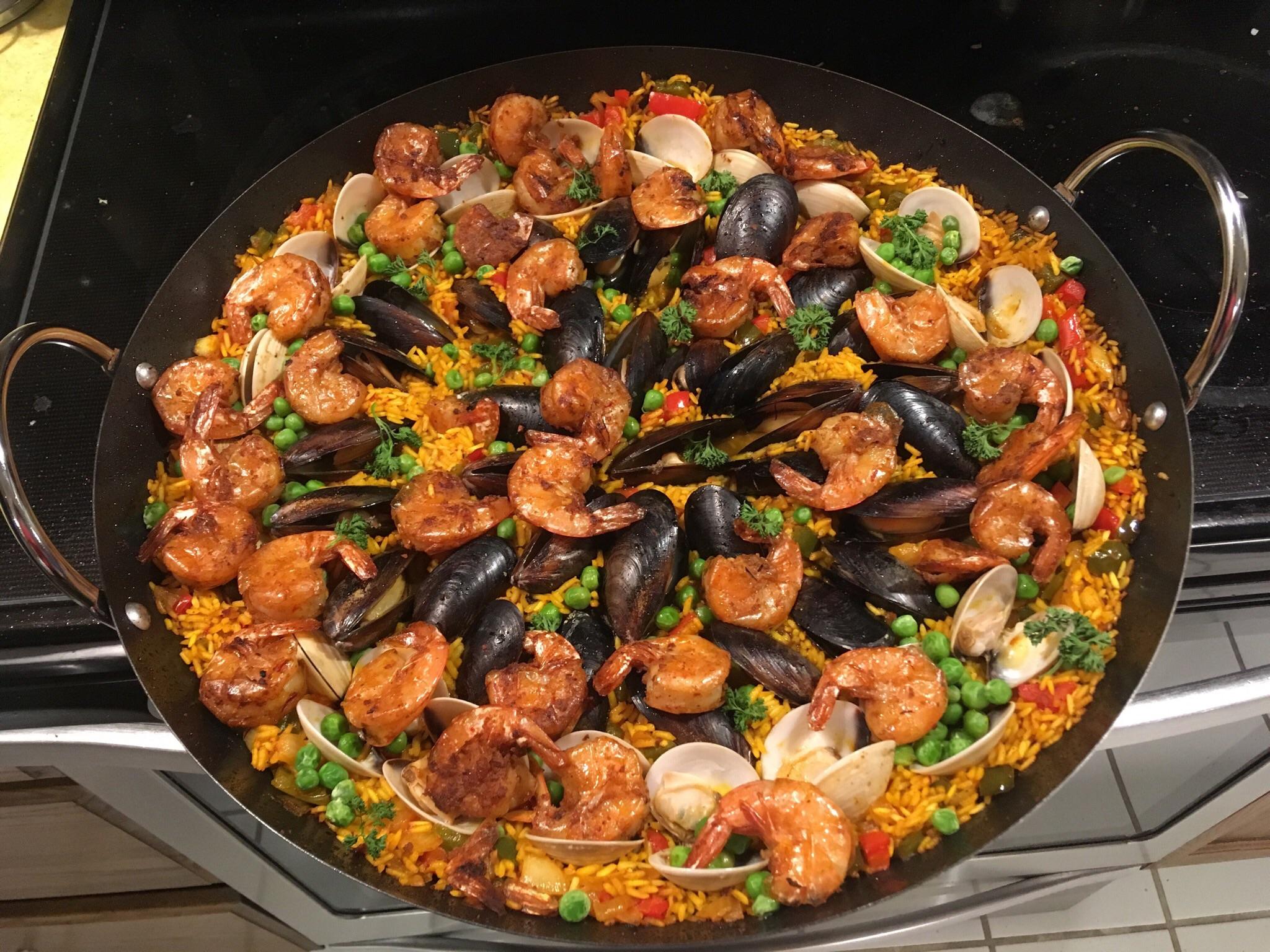 A large pan of seafood paella with shrimp, mussels, clams, peas, and red peppers on yellow rice, displayed on a stove.