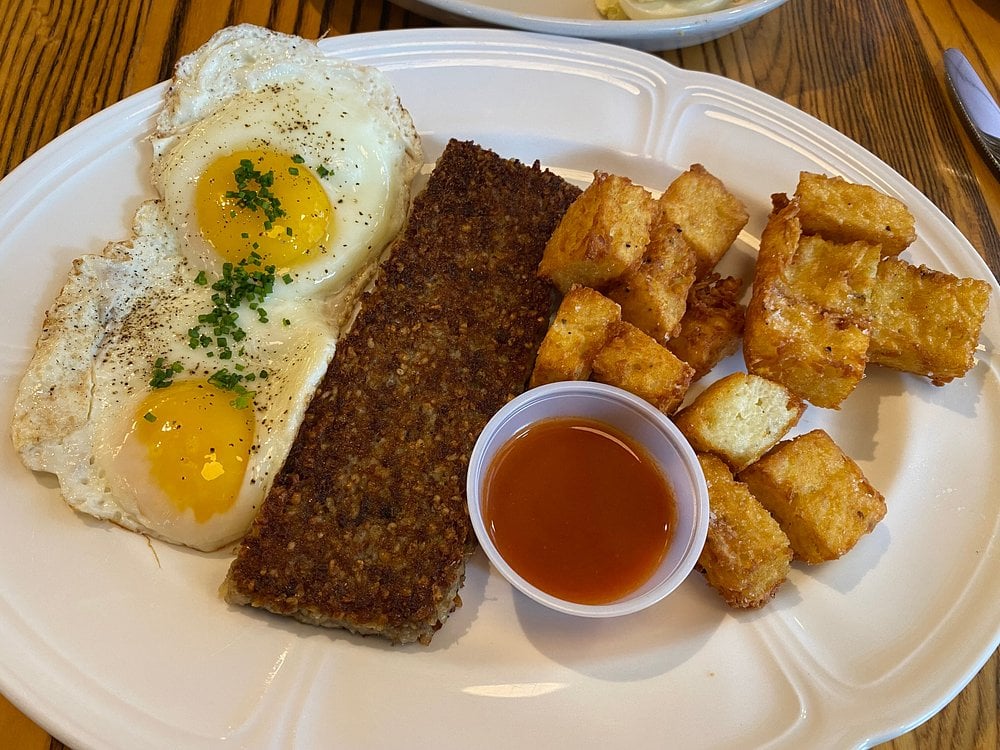 Two sunny-side-up eggs, a rectangular slice of sausage or hash, crispy golden-brown potato cubes, and a small cup of reddish dipping sauce served on a white plate.