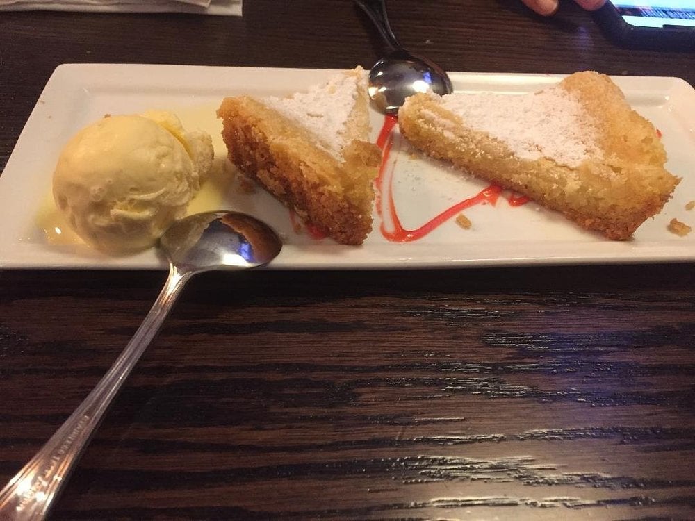 A rectangular plate with two slices of pie, a scoop of vanilla ice cream, strawberry sauce drizzle, and two metal spoons on a dark wooden table.