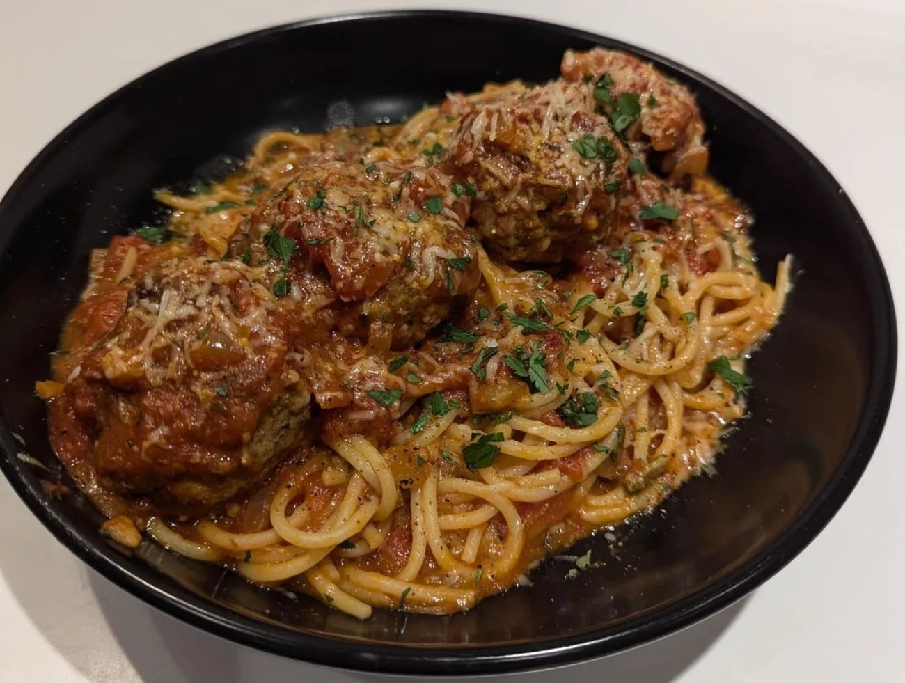 A black bowl filled with spaghetti topped with three large meatballs, marinara sauce, grated cheese, and chopped parsley, set on a white surface.