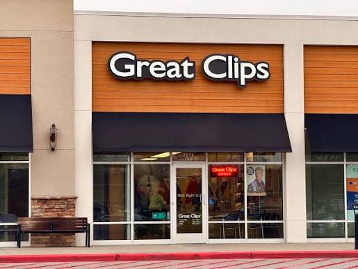 Storefront of Great Clips hair salon with a large sign above the entrance, glass doors, and windows. Some salon chairs and posters are visible inside. The exterior has orange and beige panels and black awnings.