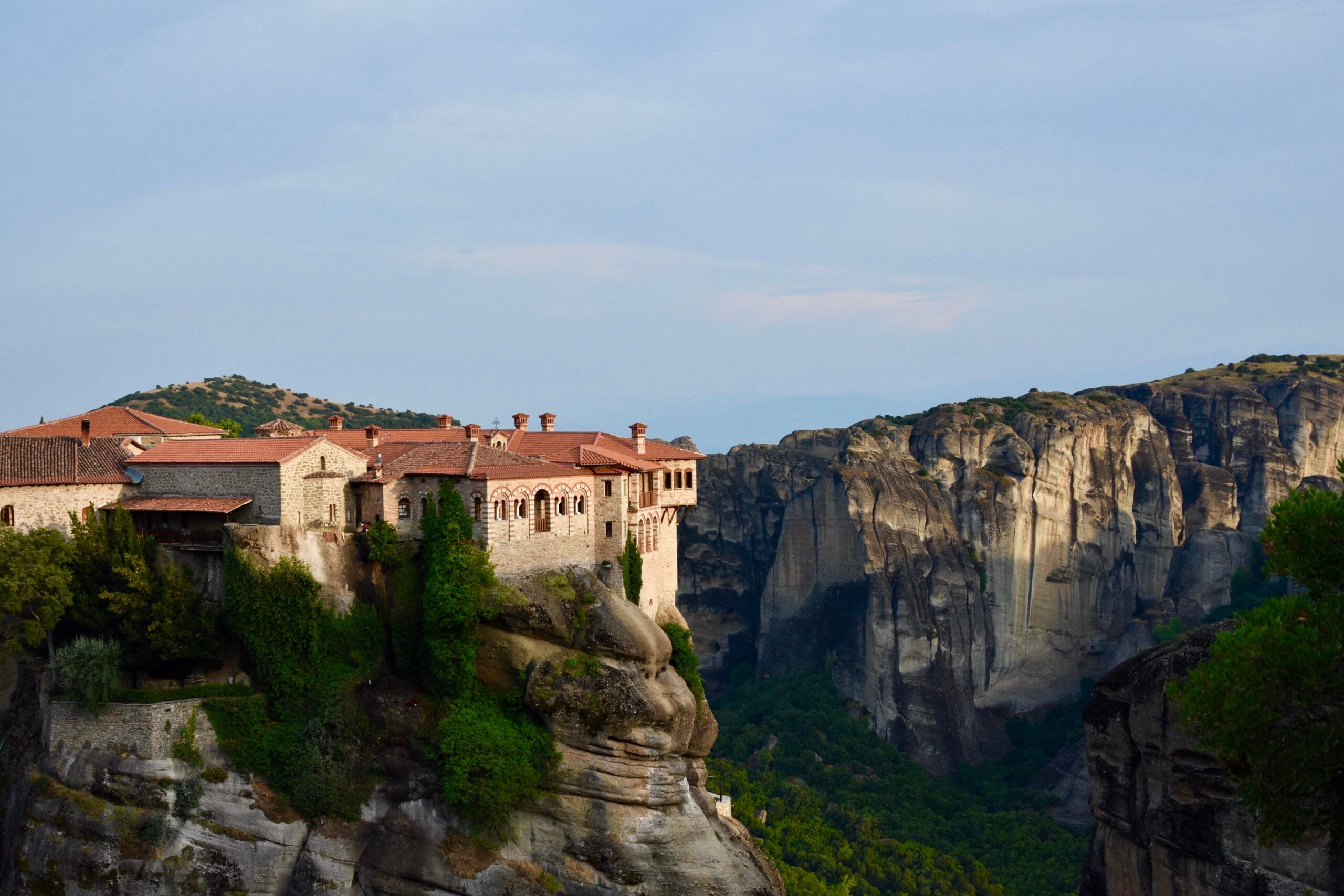 A large stone building with red roofs sits atop a steep rocky cliff surrounded by dramatic rock formations and lush green trees under a partly cloudy sky.