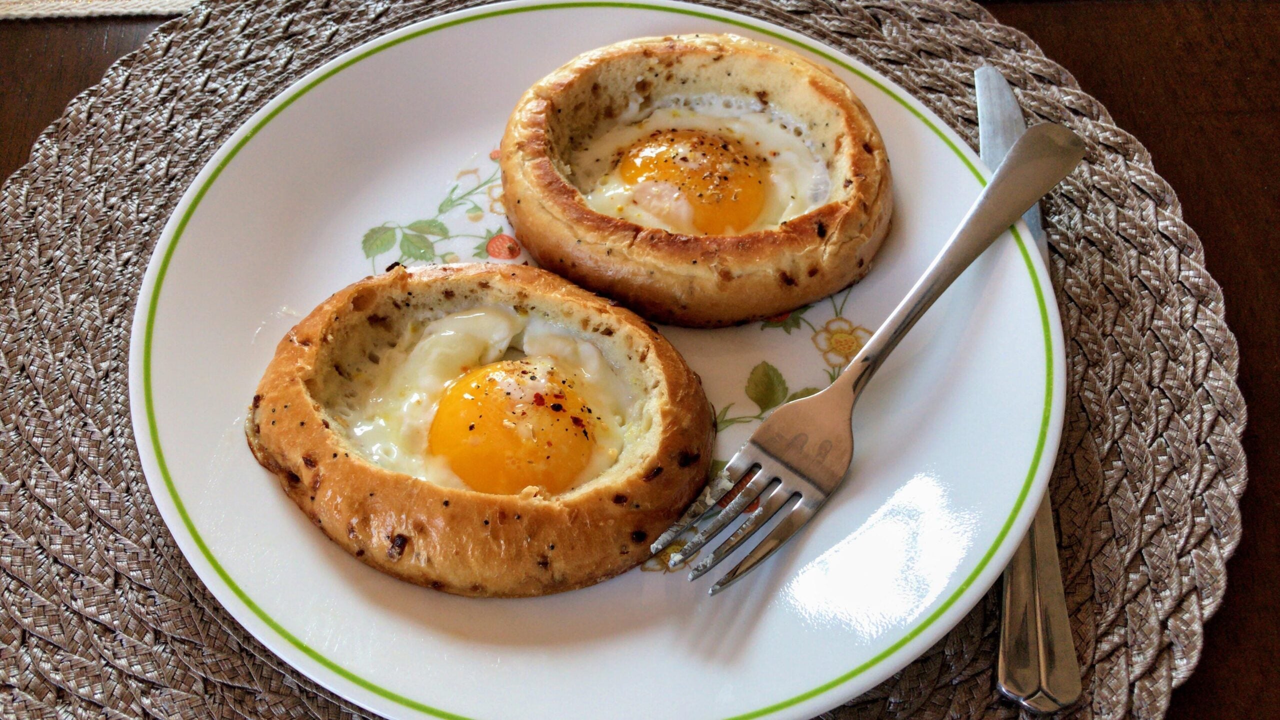 Two bagels with sunny-side-up eggs baked in the center, served on a floral plate with a fork and knife, placed on a woven placemat.