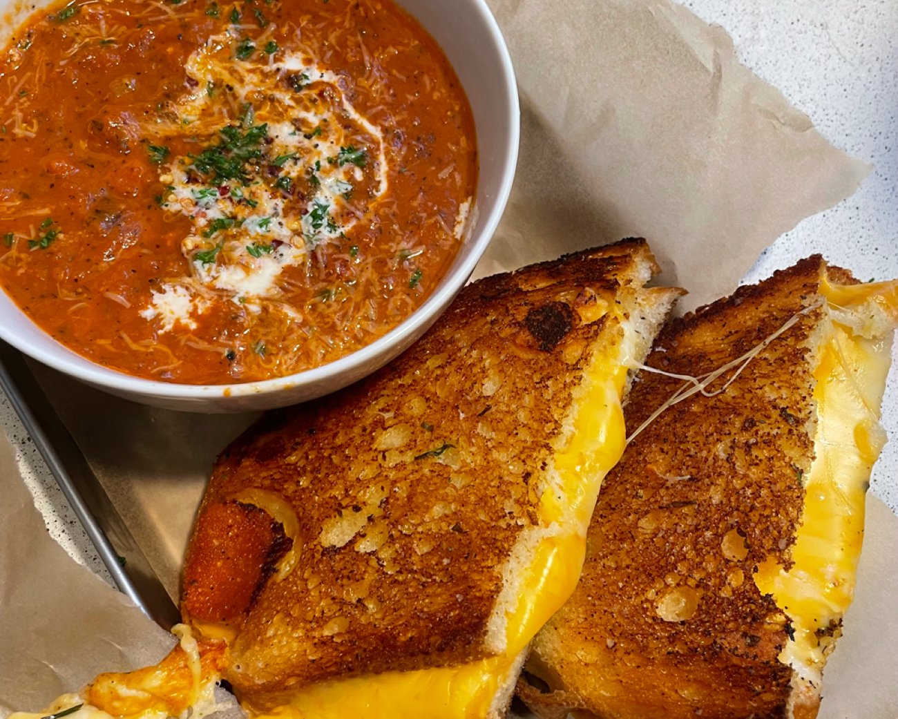 A bowl of tomato soup with herbs and cream is next to classic Grilled Cheese Sandwiches, cut in half to reveal melted yellow cheese on perfectly toasted bread.