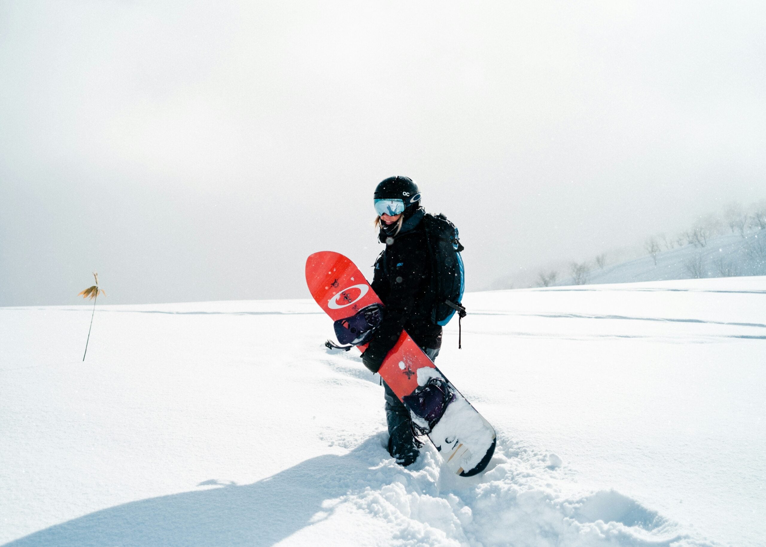 A snowboarder in black gear and goggles stands in deep snow, holding a colorful snowboard. Bright sunlight reflects off the white landscape, and distant trees are visible through the haze.