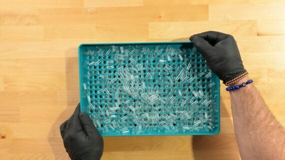 A person wearing black gloves holds a teal tray filled with small glass vials on a light wooden surface.
