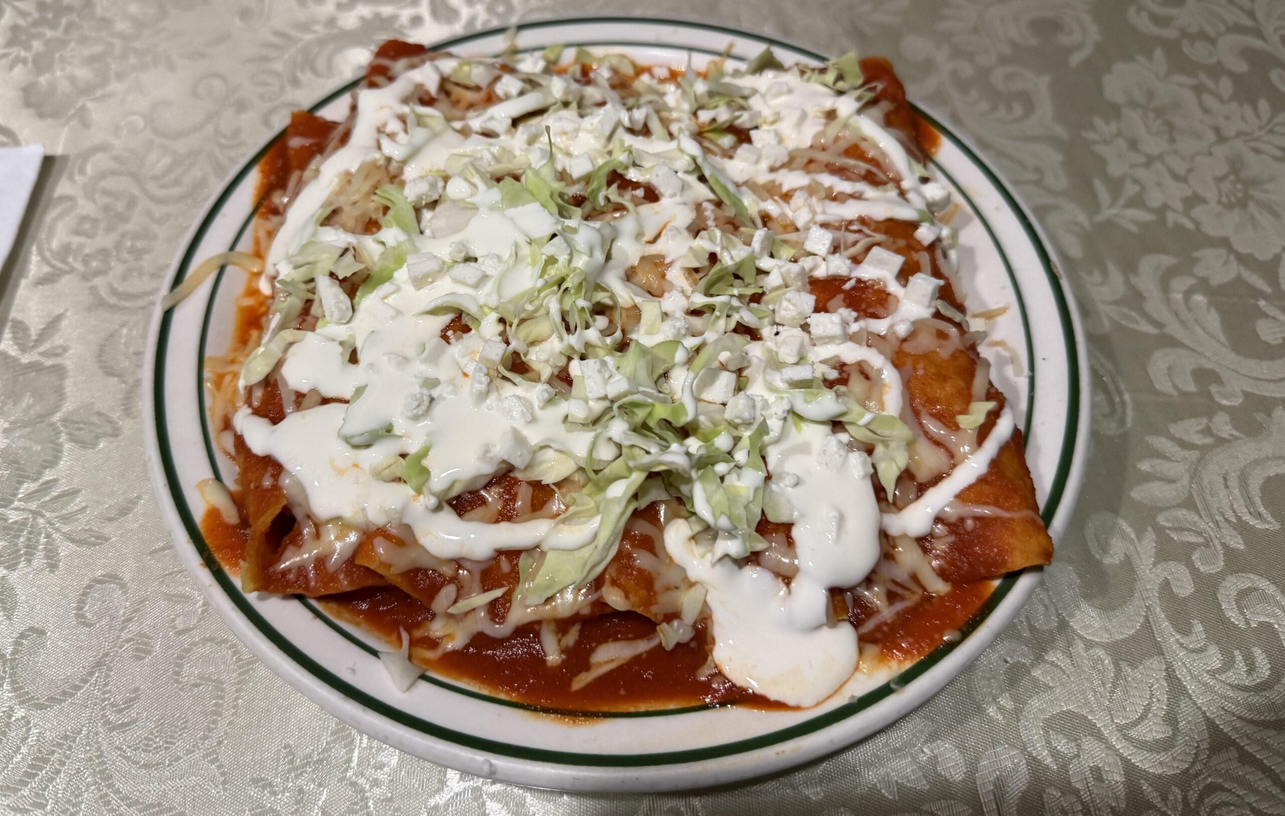 A plate of enchiladas topped with lettuce, crumbled cheese, and drizzled with sour cream sits on a patterned tablecloth.