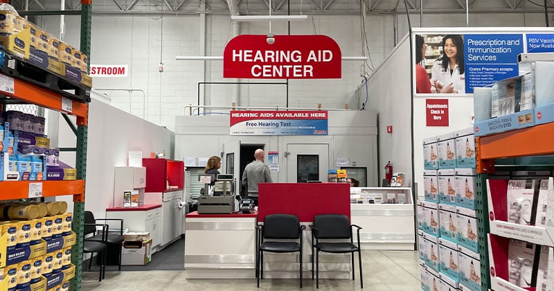 A Costco Hearing Aid Center with a red sign, desks, chairs, and shelves stocked with products; two people stand near the counter under the sign, taking advantage of free Costco membership perks.