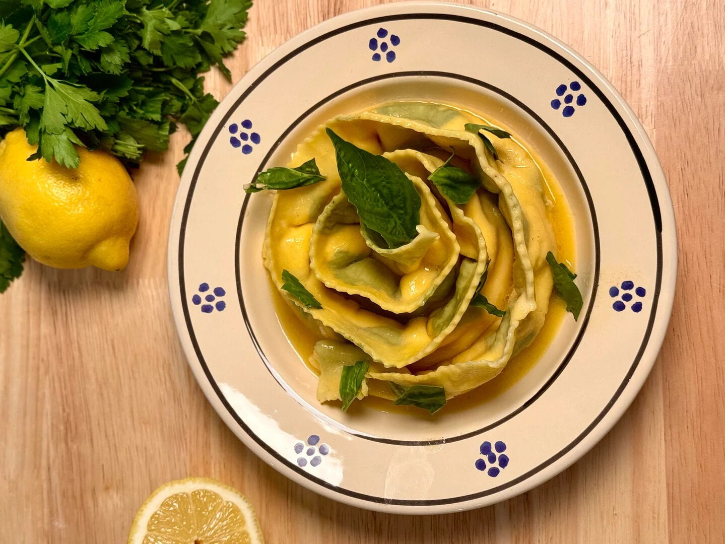 A plate of ravioli topped with fresh basil leaves sits on a wooden table, with a bunch of parsley, a whole lemon, and a lemon slice beside it. The ravioli are arranged in a swirl and served with a yellow sauce.