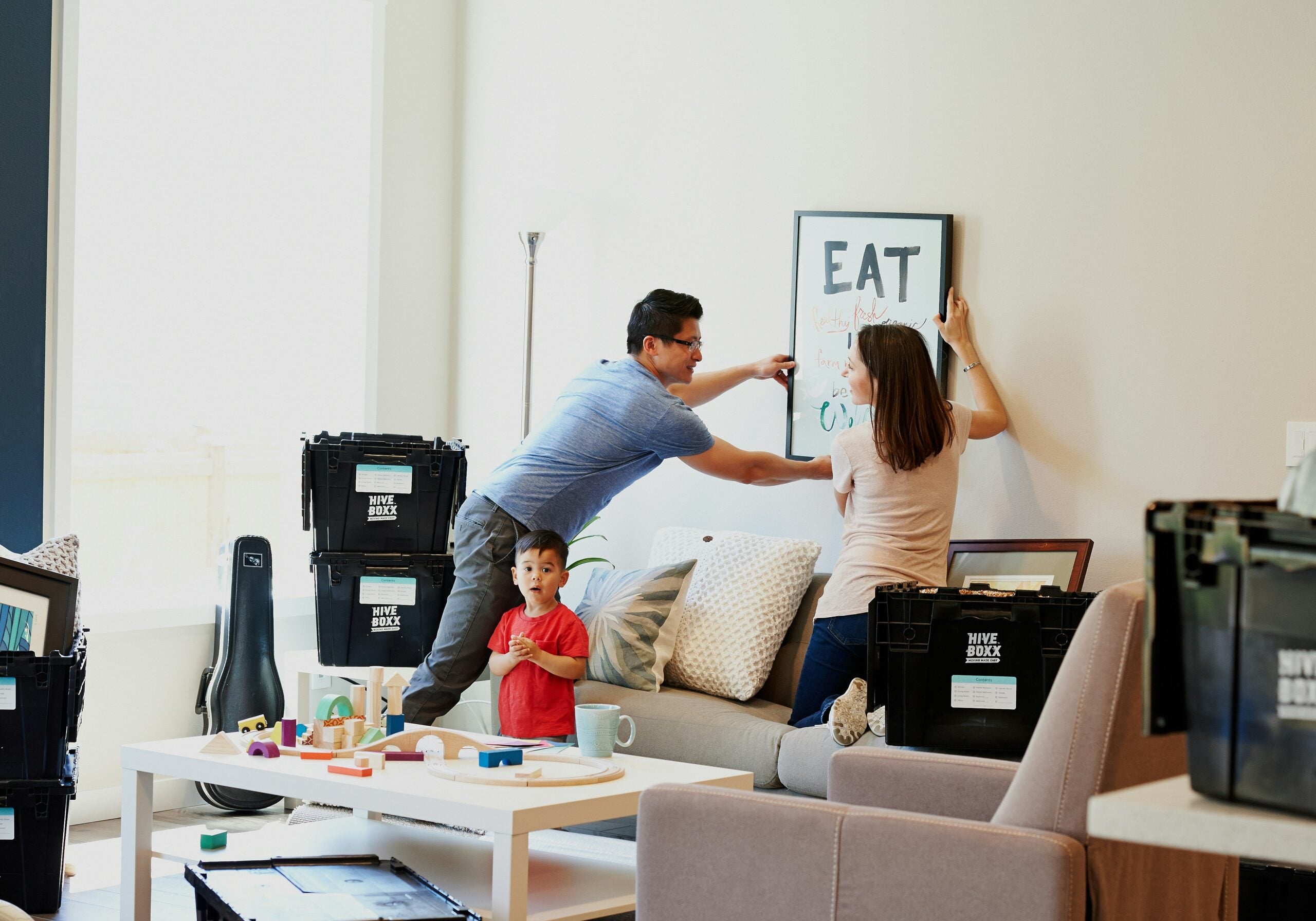 A man and woman hang a picture on the wall while a young child stands nearby. Moving boxes and toys are scattered around, suggesting the family is moving into a new home.