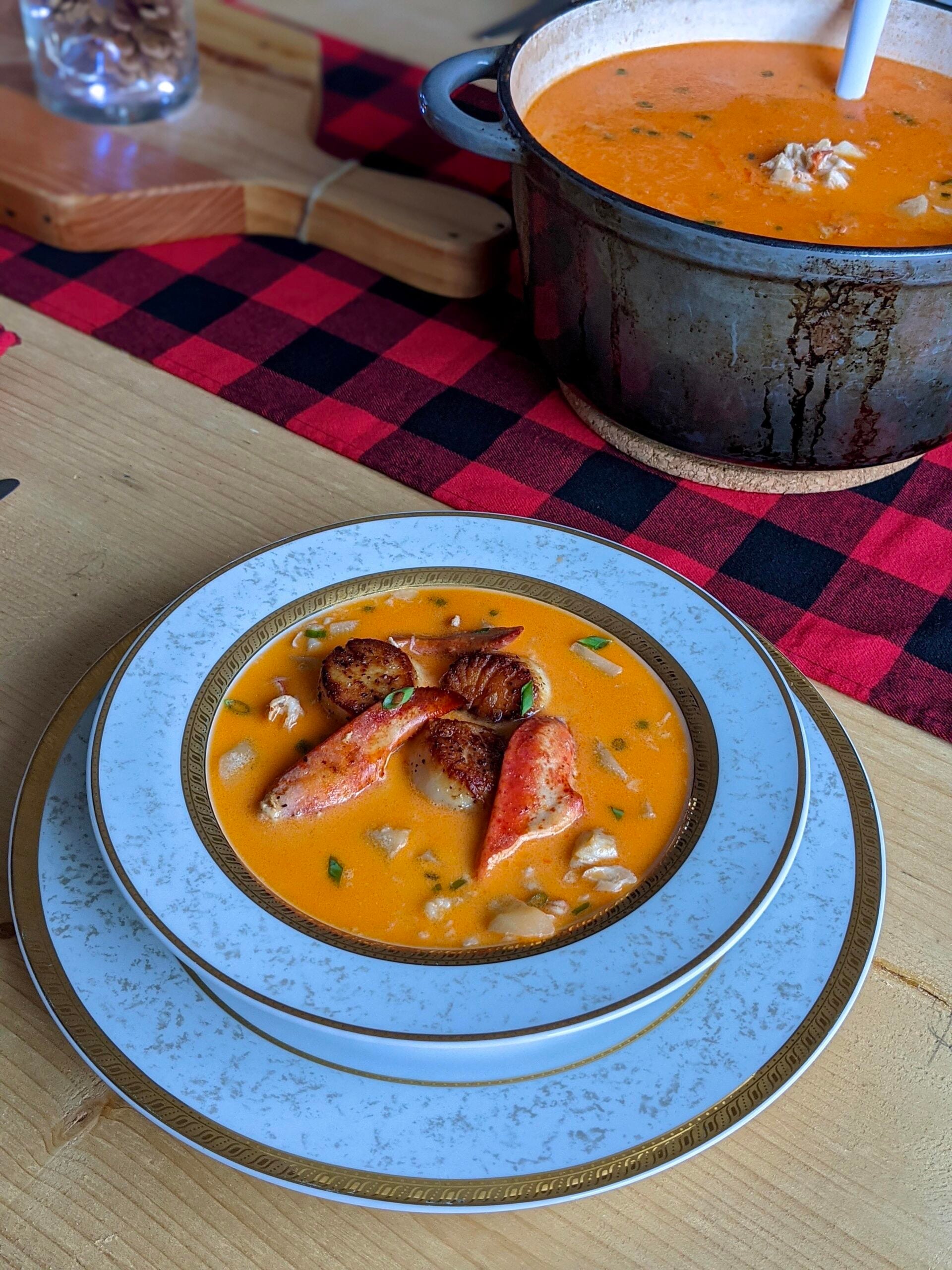 A bowl of creamy orange soup with seafood pieces is served on elegant plates. In the background, a pot of soup sits on a red and black checkered cloth atop a wooden table.