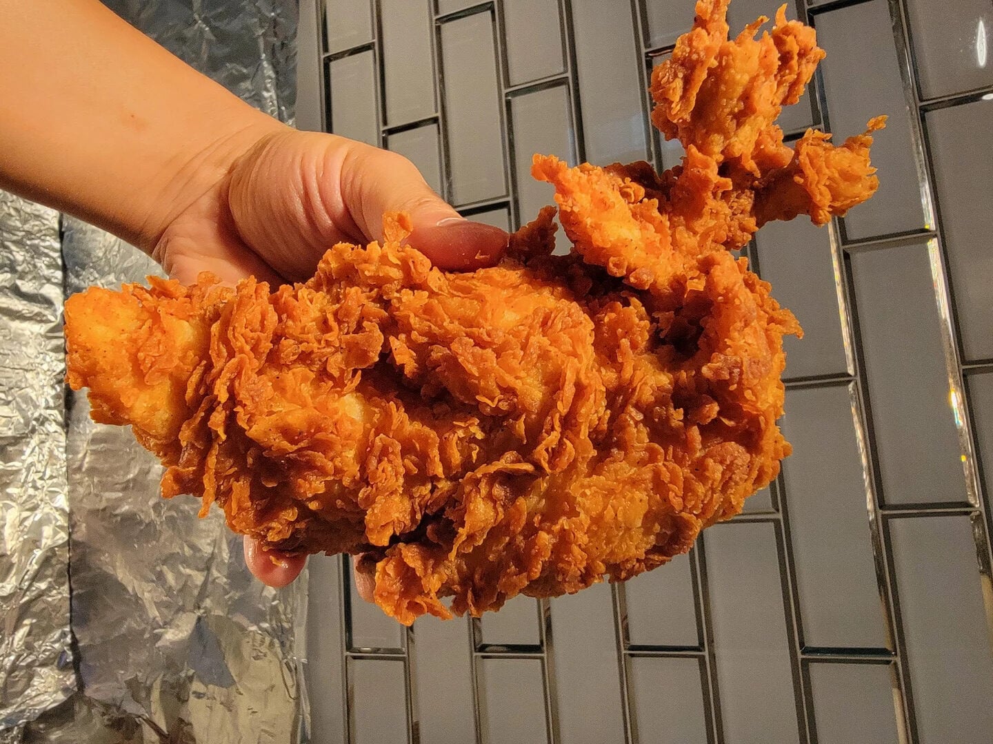 A hand holds a large, crispy piece of fried chicken in front of a tiled wall and foil-covered surface.