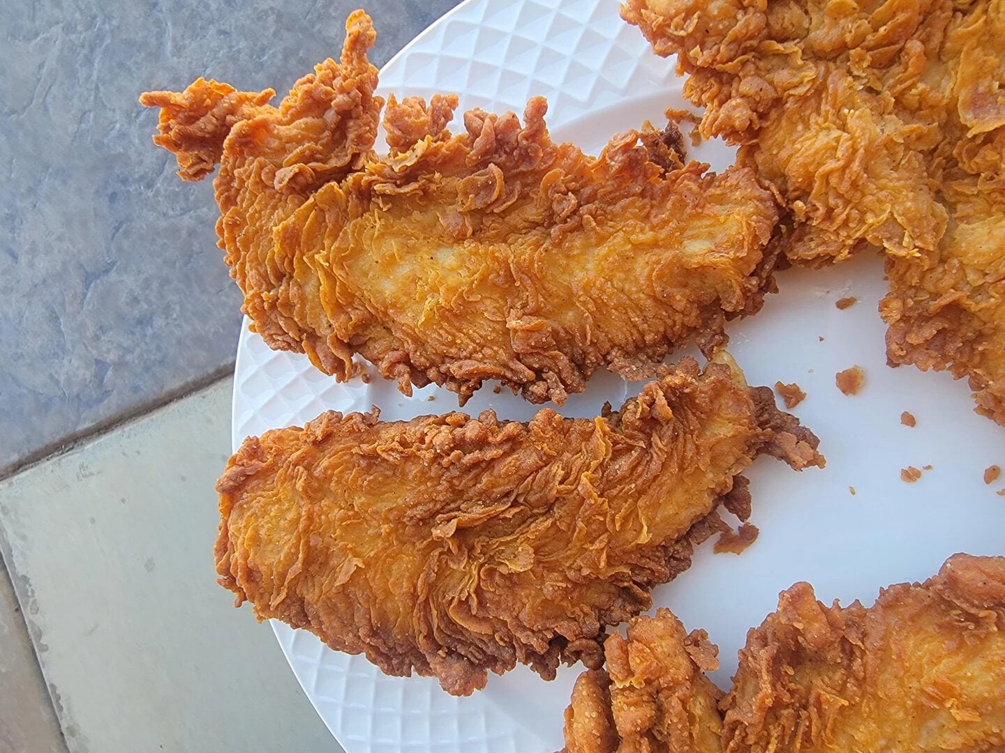 Two crispy, golden-brown fried chicken tenders on a white textured plate, with part of a gray stone surface visible in the background.