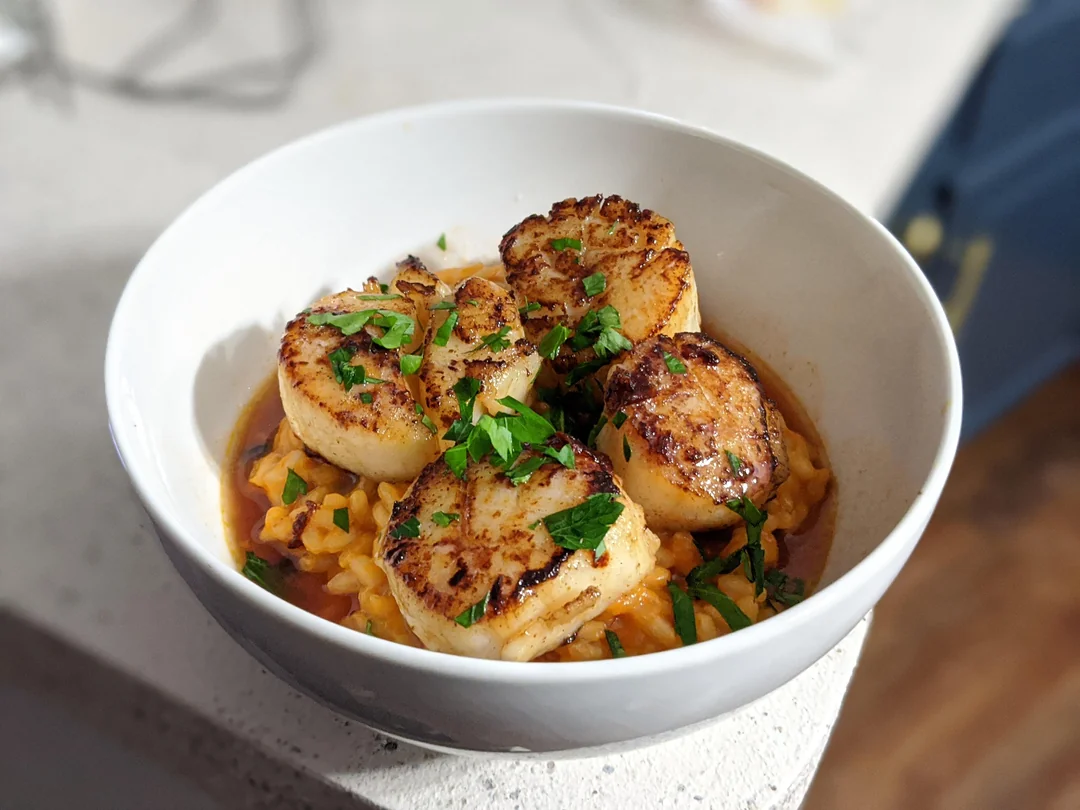 A white bowl filled with seared scallops on top of pasta in a brown sauce, garnished with fresh chopped parsley.