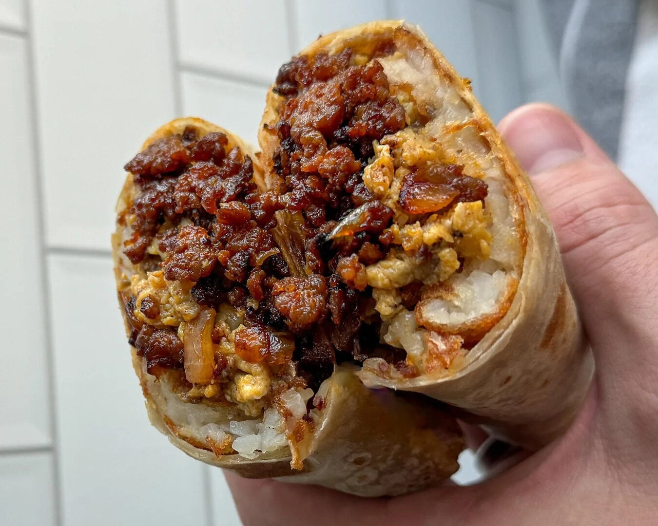 A hand holds a cut burrito filled with crispy browned meat, eggs, and potatoes, showing a savory, textured interior against a white tiled background.