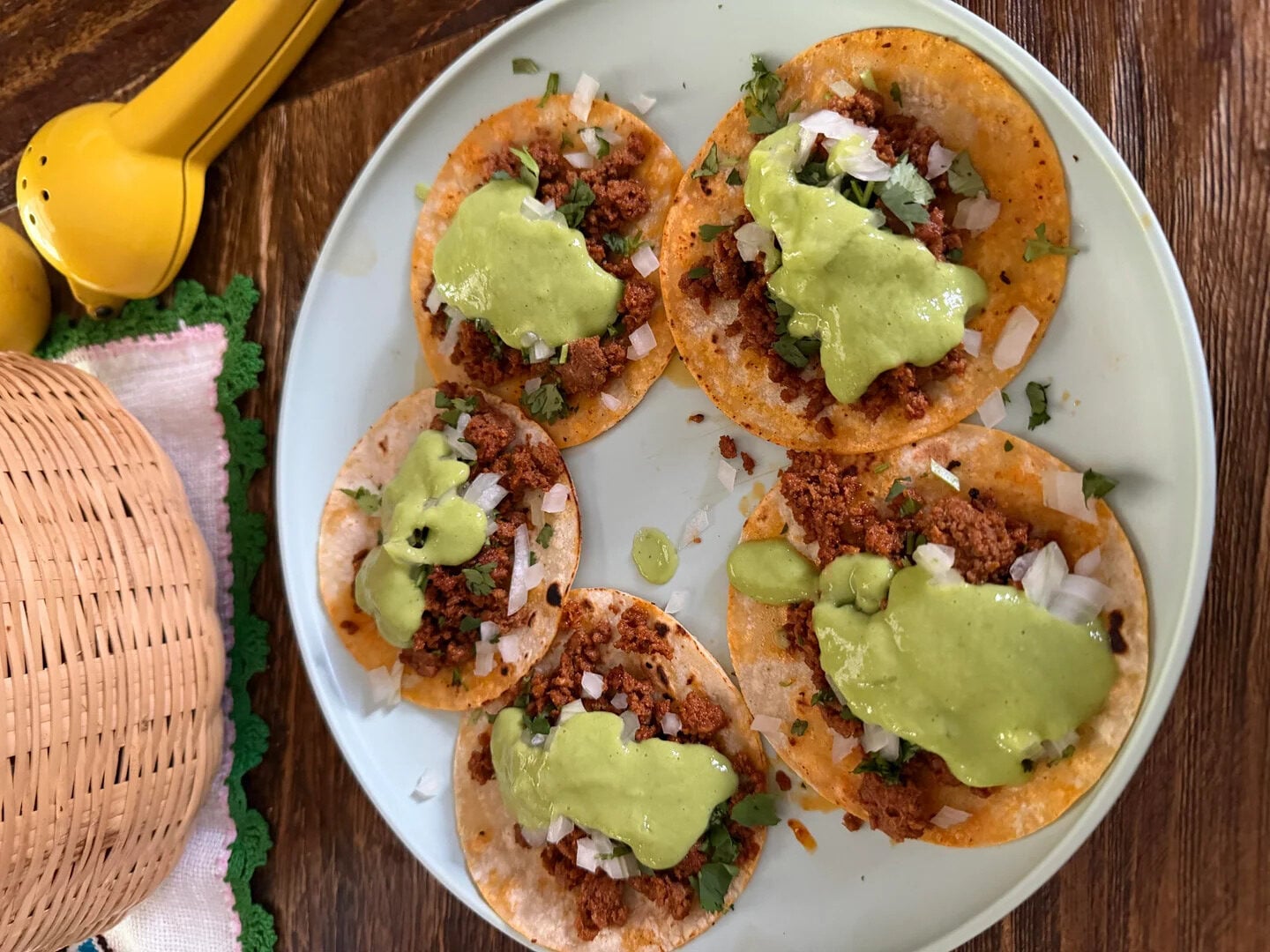 A plate of six tacos topped with chopped onions, cilantro, seasoned meat, and green sauce sits on a wooden table next to a yellow lemon squeezer and a woven basket.