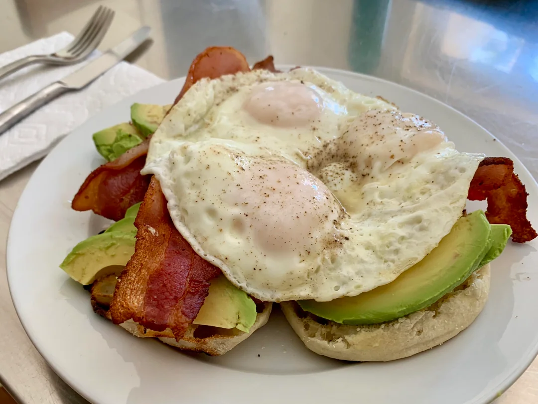 Two fried eggs on top of sliced avocado, crispy bacon, and an English muffin, served on a white plate with a fork and knife on a napkin beside it.