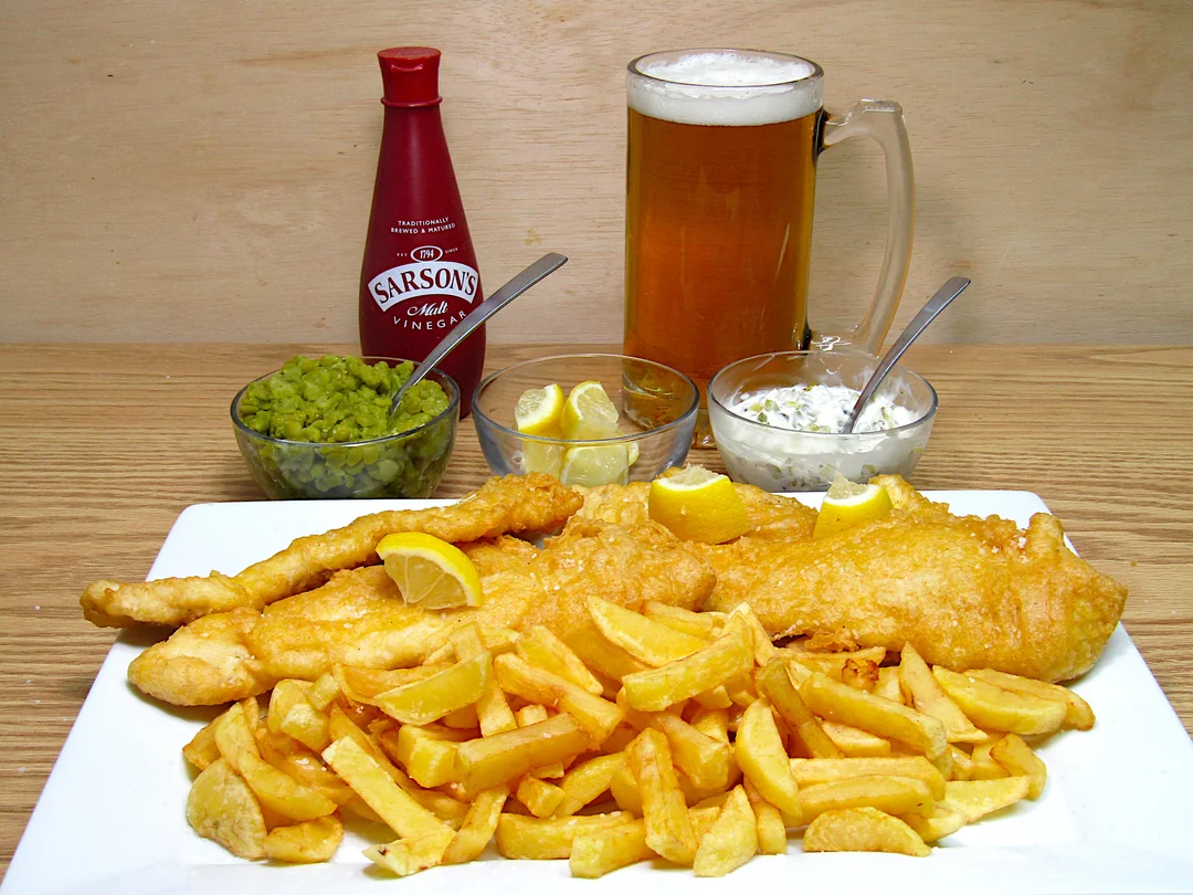 A plate of fish and chips with lemon wedges, mushy peas, tartar sauce, and a mug of beer. A bottle of Sarson’s vinegar is in the background.