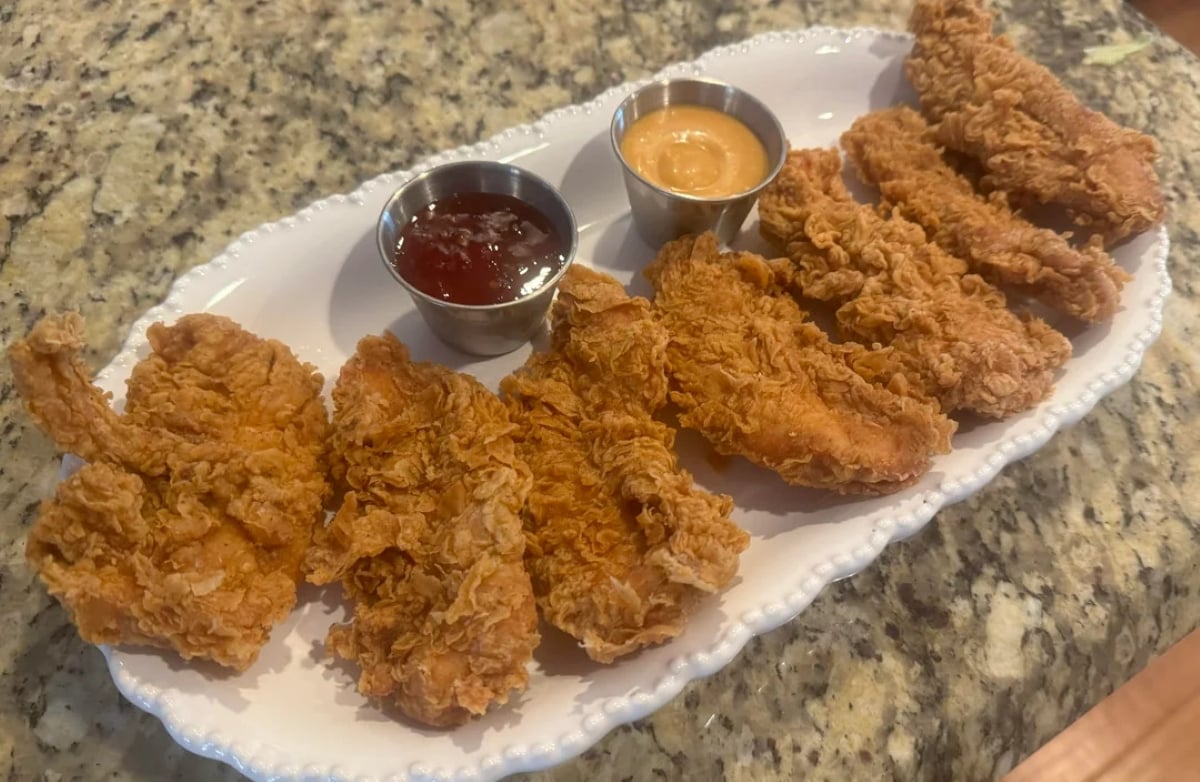 A white plate holds eight crispy fried chicken tenders with metal cups of red and orange dipping sauces, set on a granite countertop.