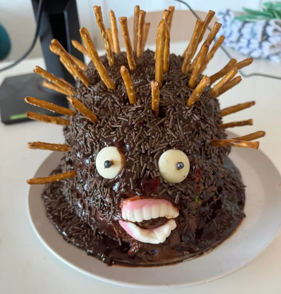 A round cake covered in chocolate sprinkles and pretzel sticks, resembling a hedgehog. It has candy eyes and large candy teeth, giving it a quirky and funny appearance.