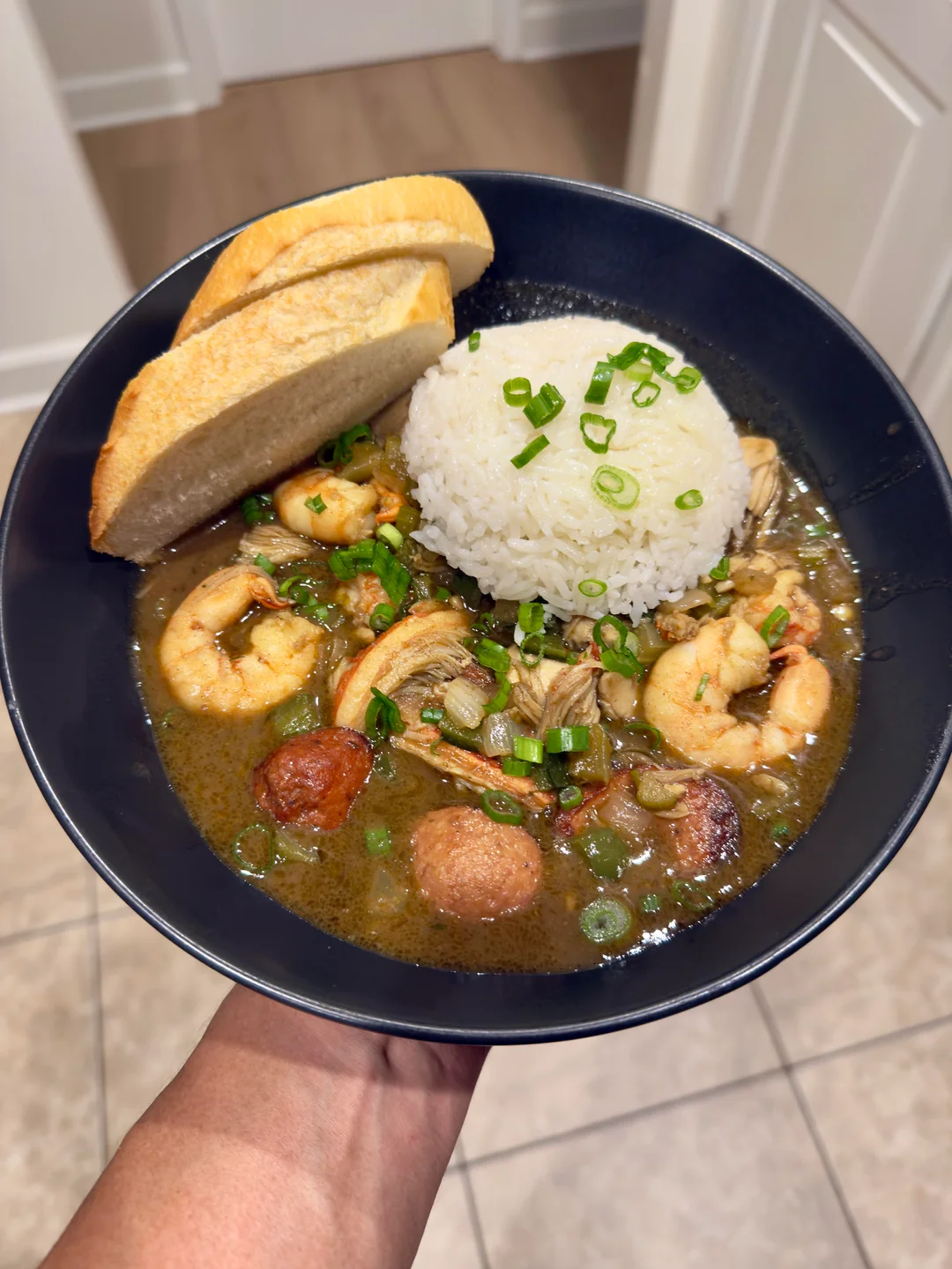 A bowl of gumbo with shrimp, sausage, and vegetables, topped with green onions, served with a dome of white rice and two slices of bread.