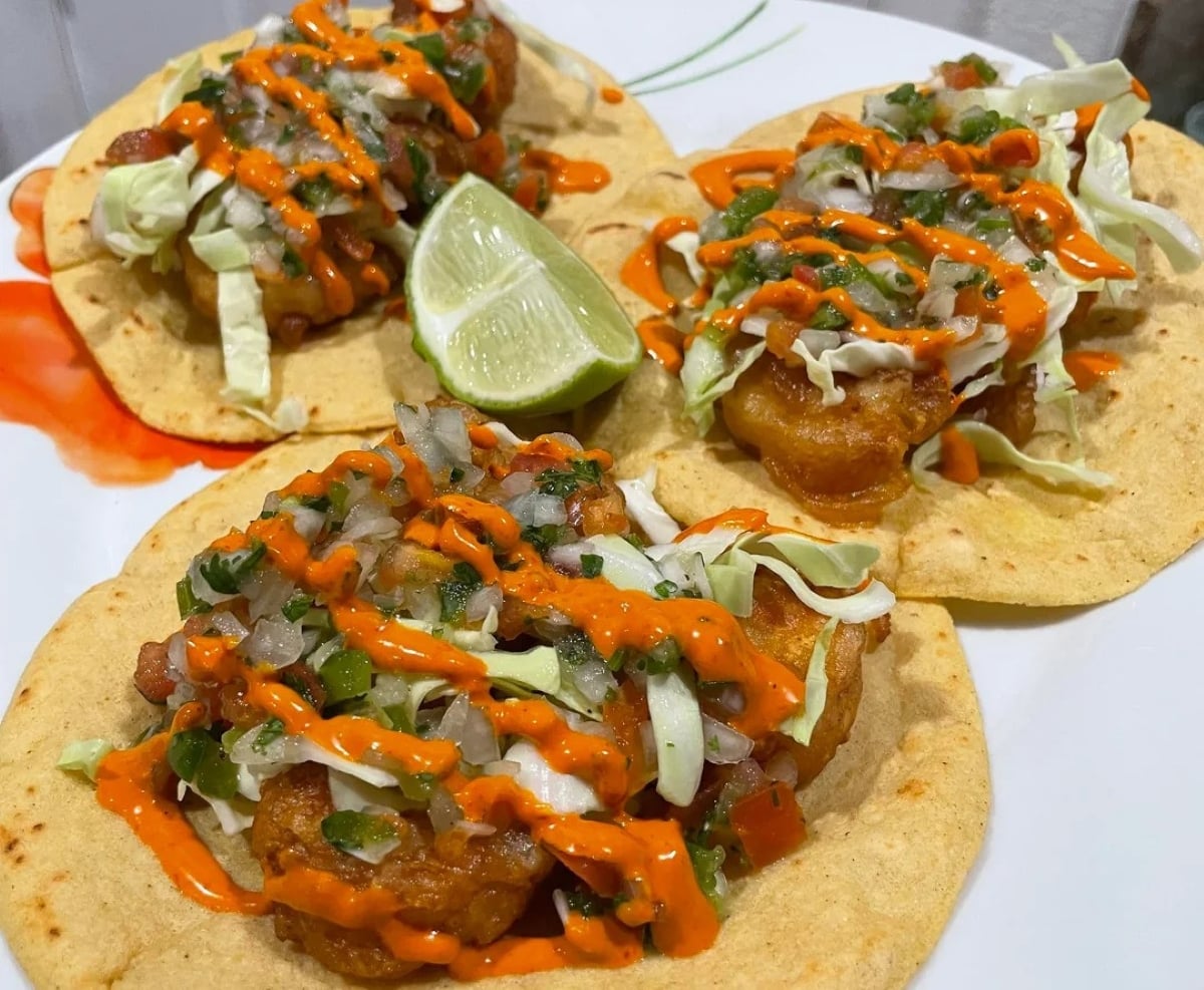 Three shrimp tacos topped with chopped cabbage, salsa, and orange sauce are served on corn tortillas with a lime wedge on the side.