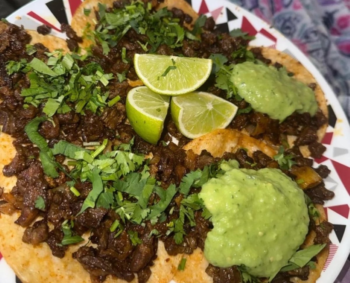 A plate of tacos topped with chopped beef, fresh cilantro, two lime wedges, and dollops of green salsa on a colorful patterned dish.