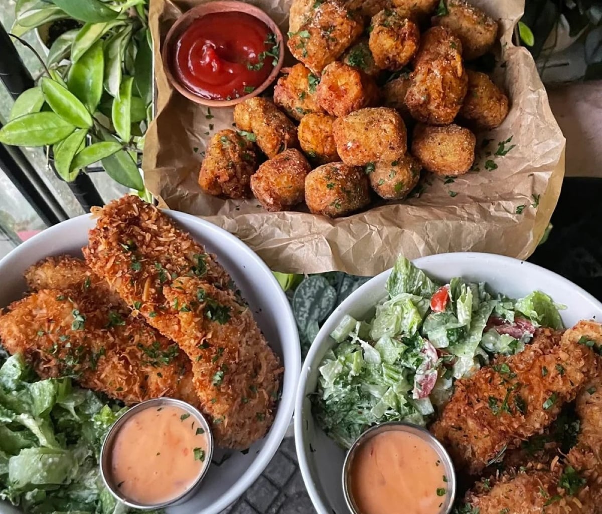 Two bowls of salad with breaded, fried pieces and dipping sauce, next to a basket of seasoned tater tots with ketchup, all garnished with herbs.