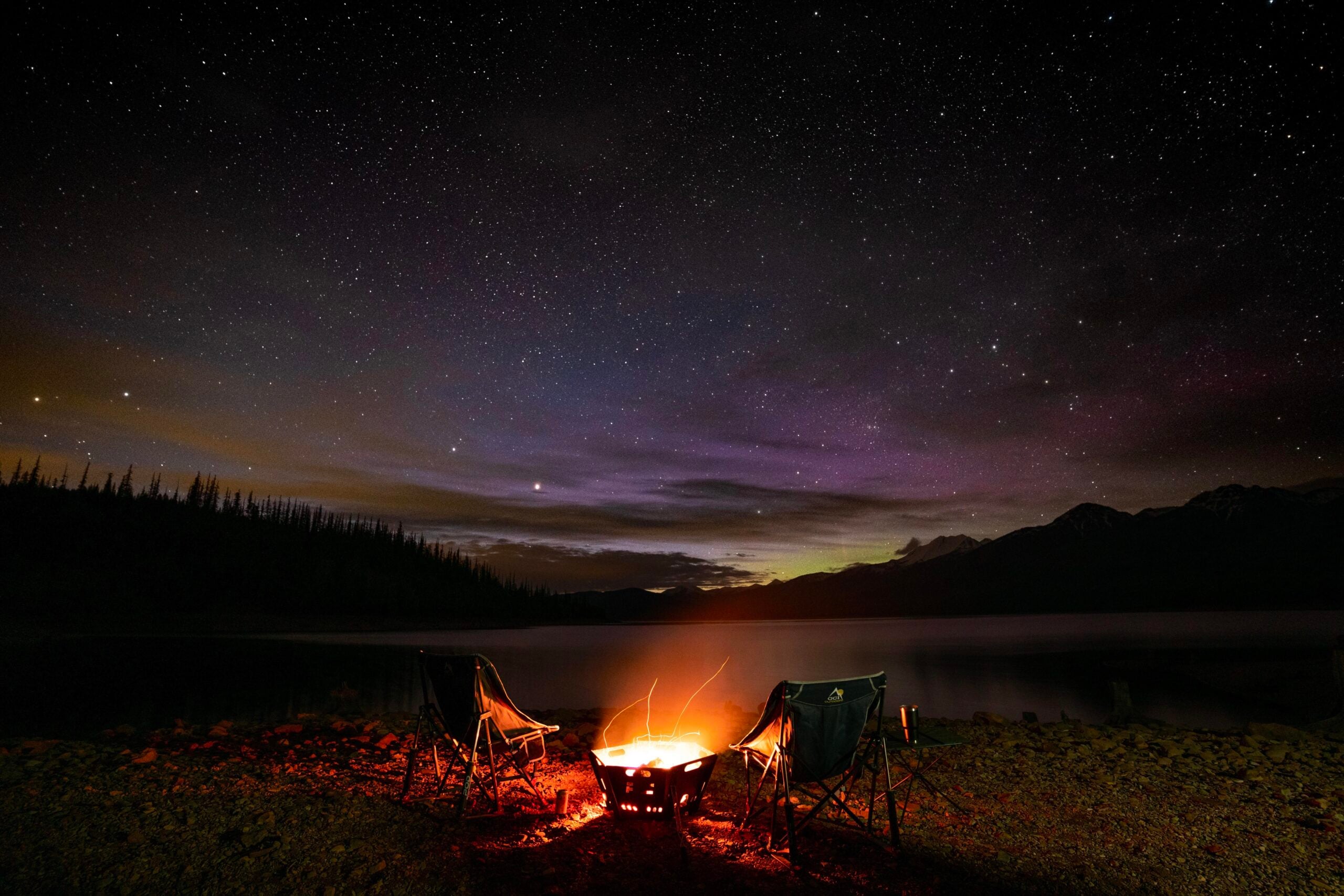 Two empty camping chairs face a glowing campfire by a lake under a star-filled night sky, with mountains and trees silhouetted in the background.