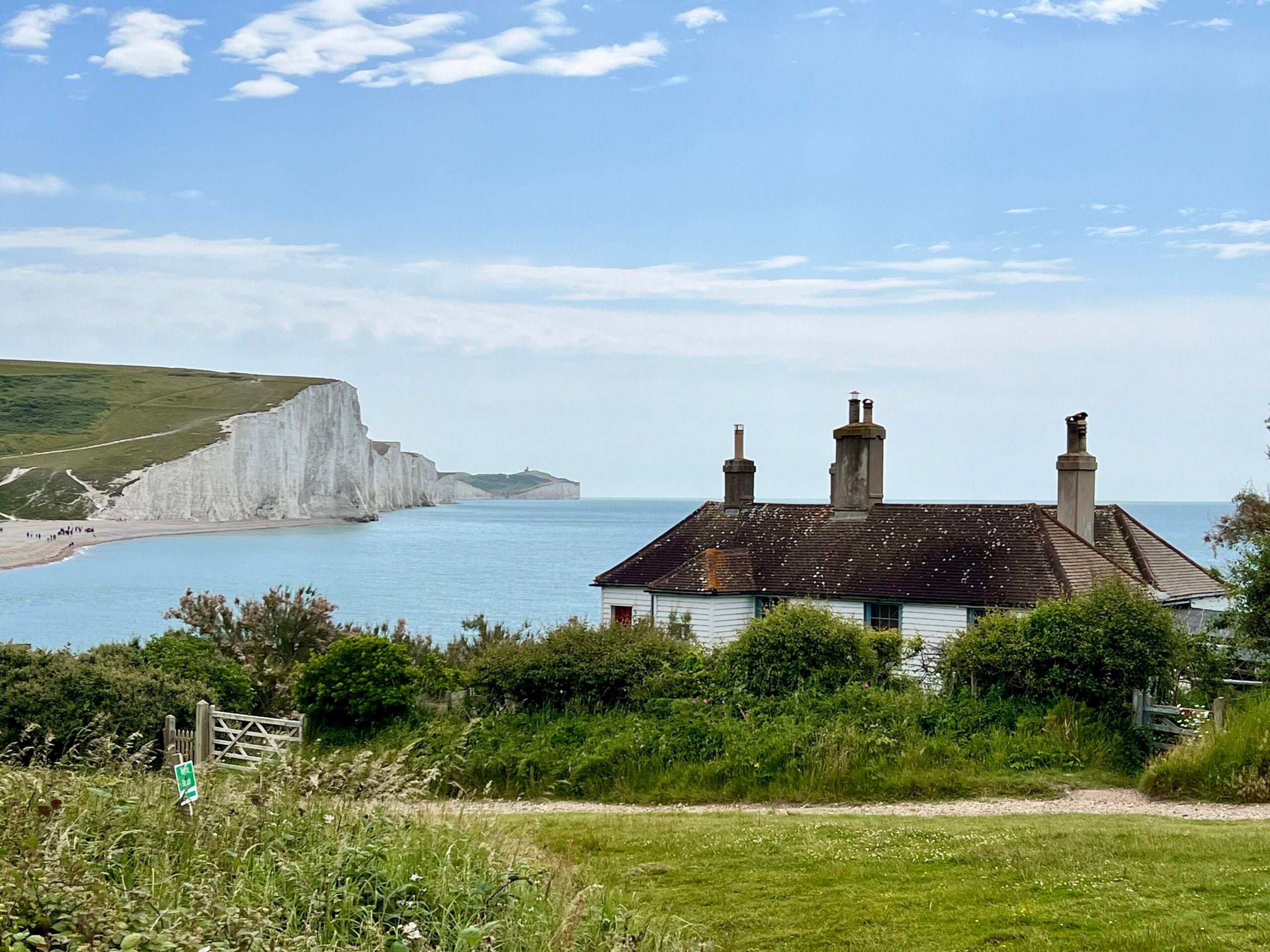A small cottage with chimneys sits by the sea, overlooking dramatic white chalk cliffs and a calm blue coastline under a partly cloudy sky. Grassy plants and a wooden gate are visible in the foreground, all set just steps away from some of the region’s incredible beaches.