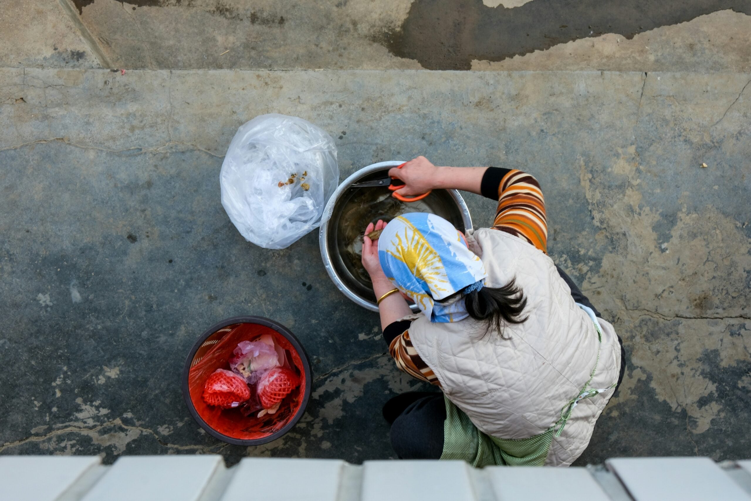 A person wearing a headscarf and striped sleeves washes food in a large metal bowl, with two bins nearby, one for trash and one for food scraps, on a concrete floor.