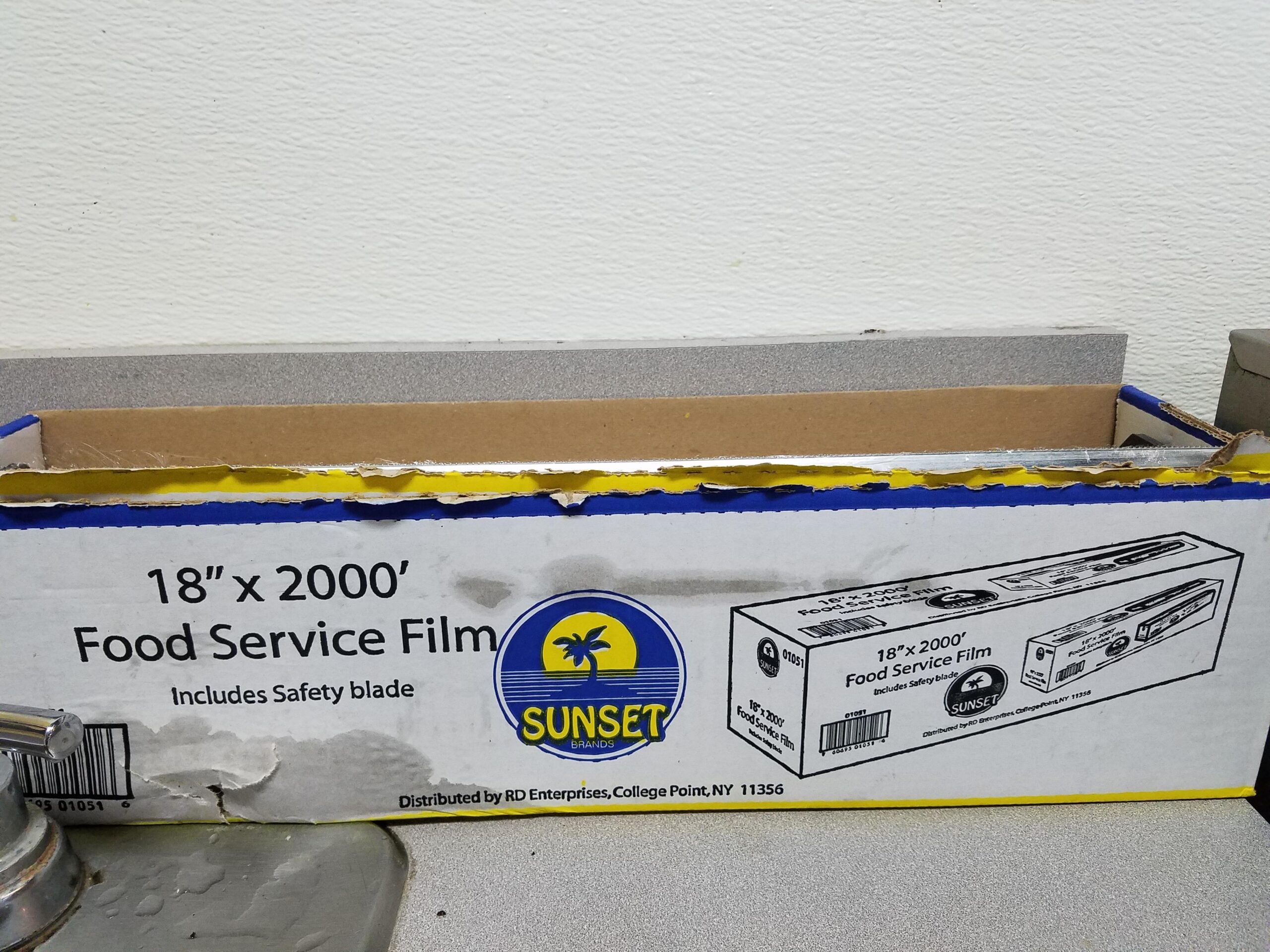 A box of Sunset brand 18″ x 2000′ food service film sits on a gray countertop, partially opened. The box is white with blue and yellow accents and includes a safety blade.