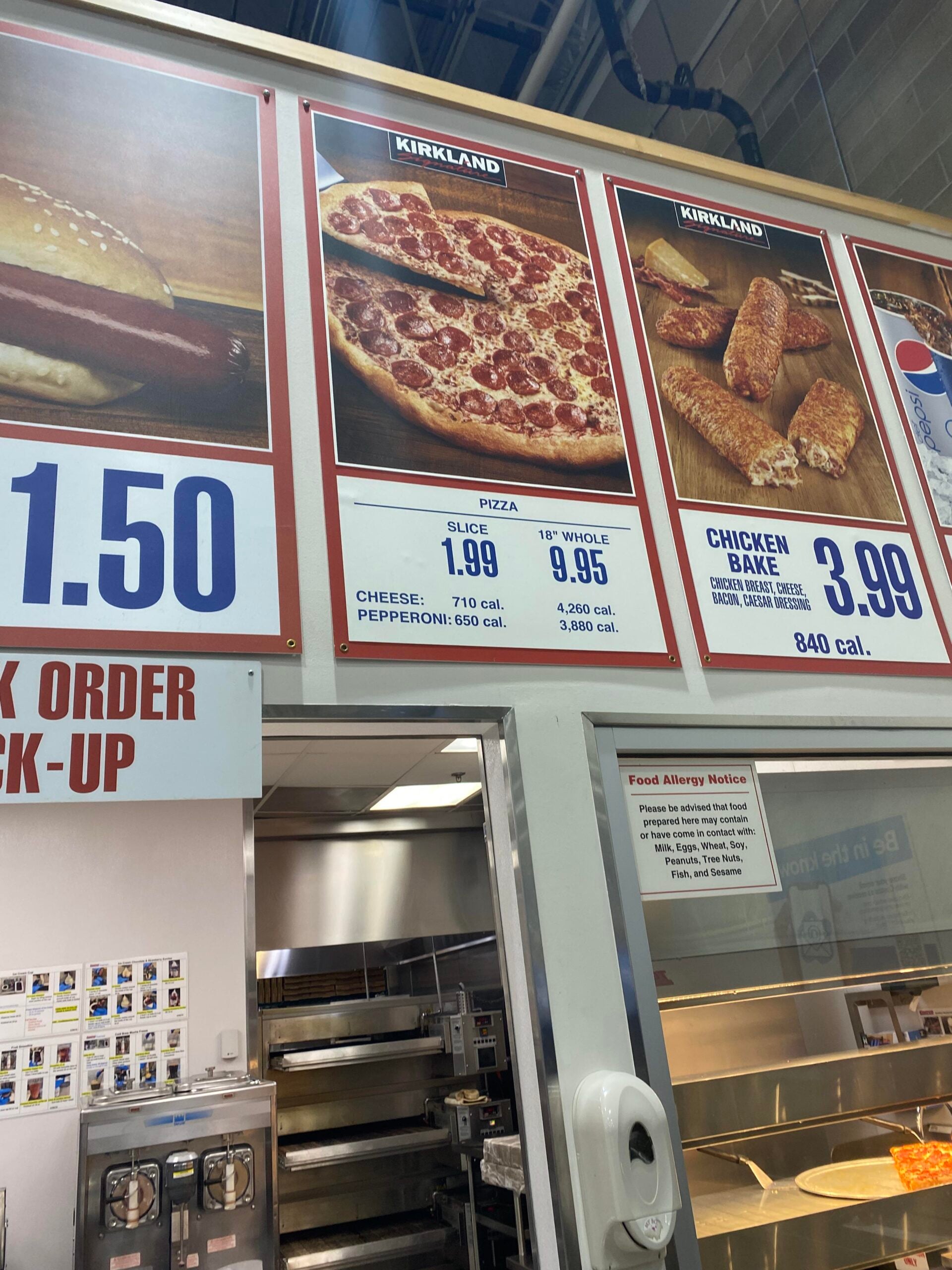Menu board above a counter shows prices for Kirkland pizza by the slice or whole, chicken bake, and a hot dog combo. Calorie counts are listed under each item. Part of the kitchen and food pickup area is visible below.