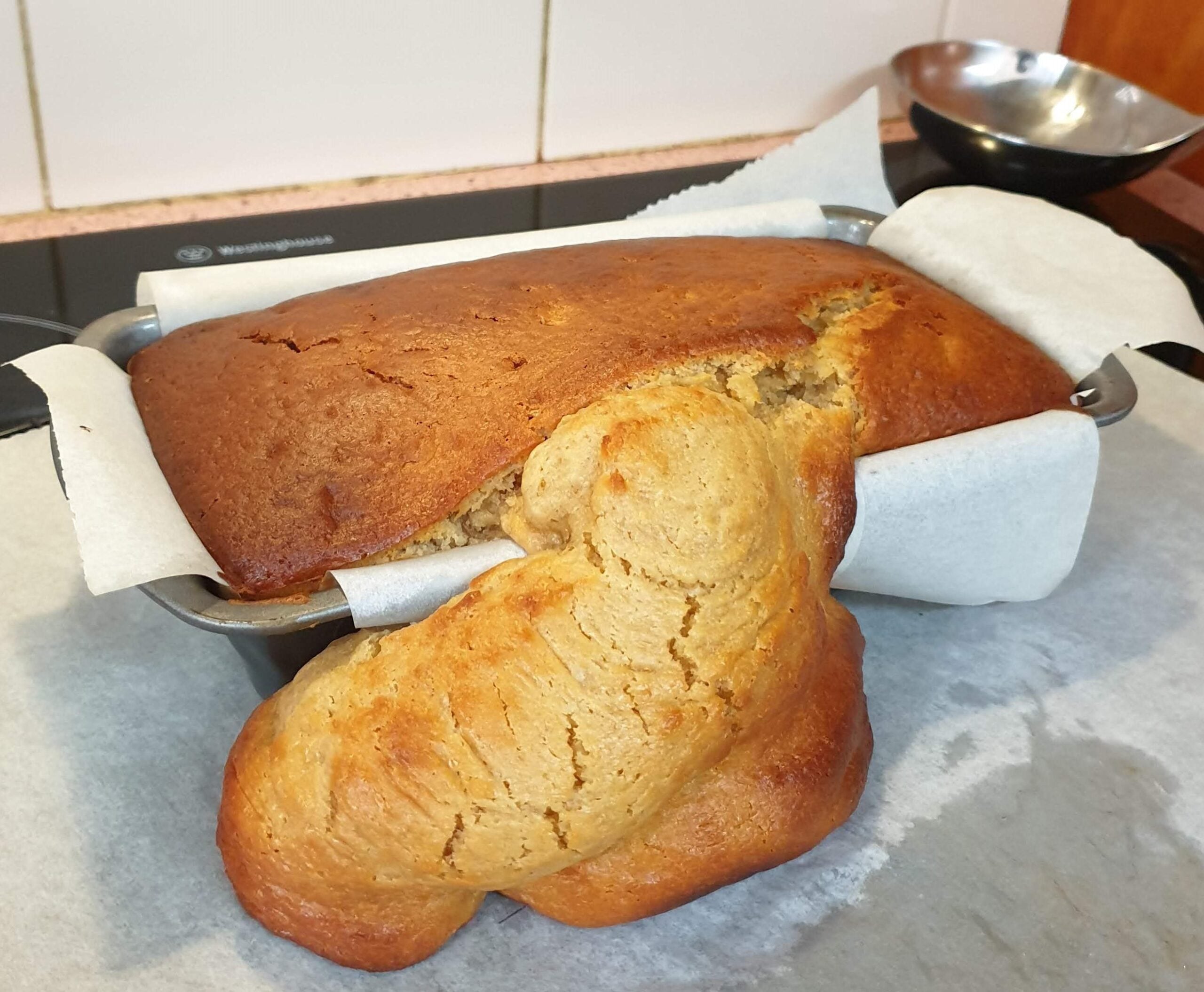 Banana bread overflowing from a loaf pan, with a large, oddly shaped portion baked onto the side, resting on parchment paper on a countertop.
