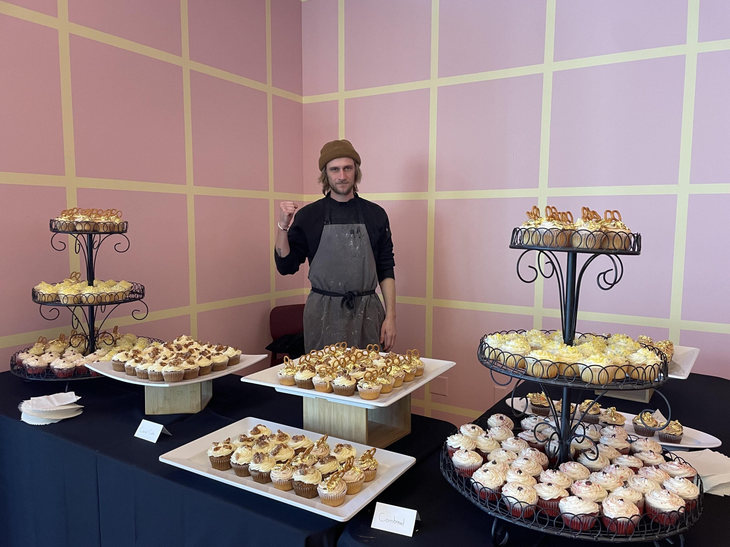 A person in an apron and hat stands behind a table filled with assorted cupcakes on trays and tiered stands, against a pink-and-yellow checkered wall.