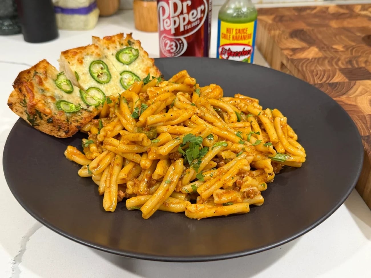 A black plate of pasta with sauce and herbs, served with two slices of jalapeño-topped garlic bread. Dr Pepper and a hot sauce bottle are in the background on a kitchen counter, making for mouthwatering photos of pasta that capture both flavor and ambiance.
