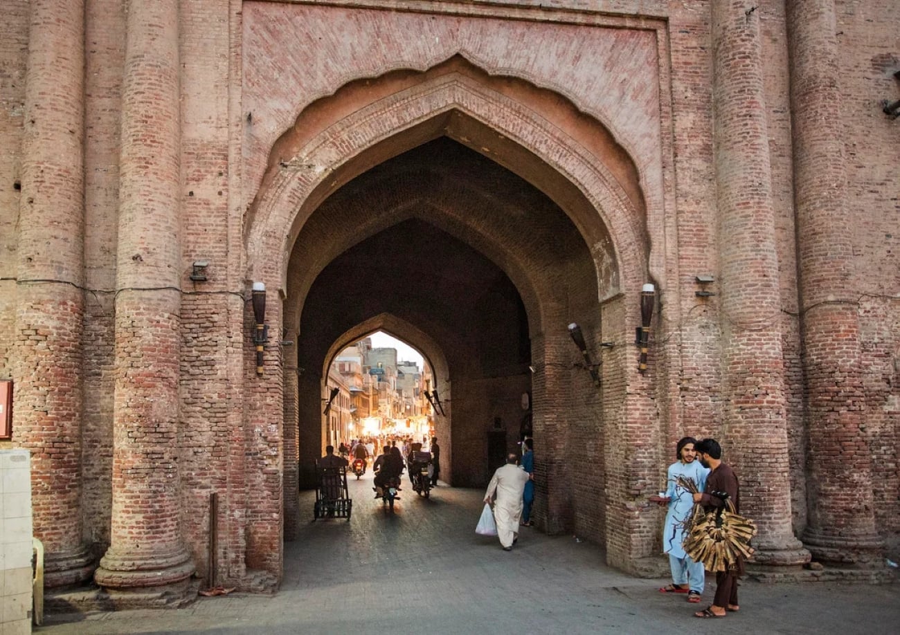 A large brick archway serves as an entrance to a bustling street in one of the Biggest Cities In The World, with people walking, a man carrying firewood, and lights visible inside. The scene depicts a lively urban area at dusk.