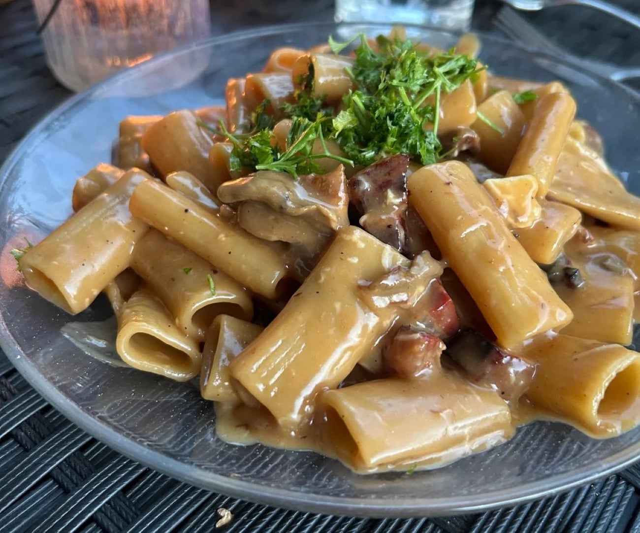 A glass plate of creamy pasta with mushrooms and herbs, garnished with fresh parsley, sits on a woven black placemat—one of many inviting photos of pasta that capture both flavor and elegance.