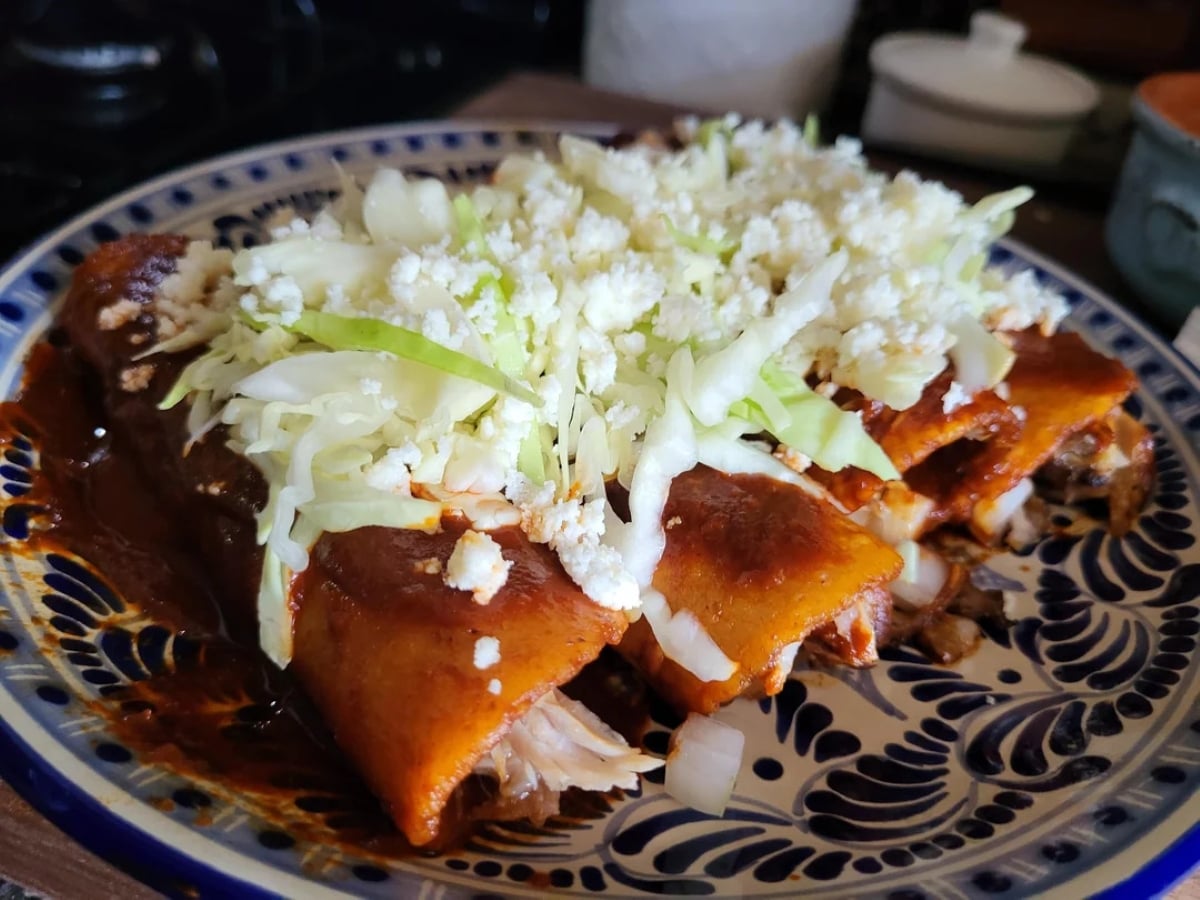 Three enchiladas topped with chopped lettuce, onions, and crumbled cheese on a decorative ceramic plate. The enchiladas are covered in red sauce and appear filled with shredded meat.