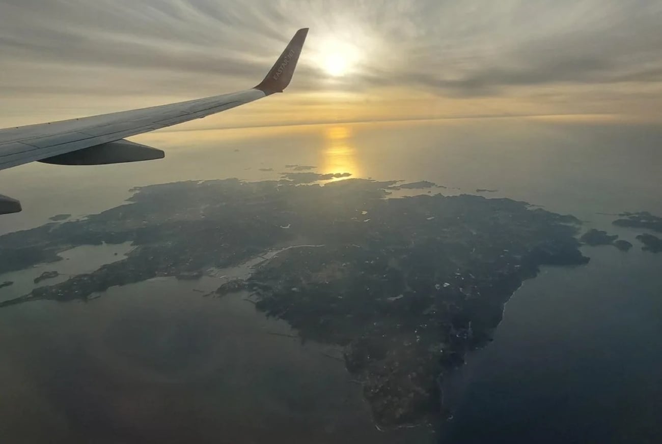 View from airplane windows showing the wing, cloudy sky, and sunset reflecting over a large island and the surrounding water below.