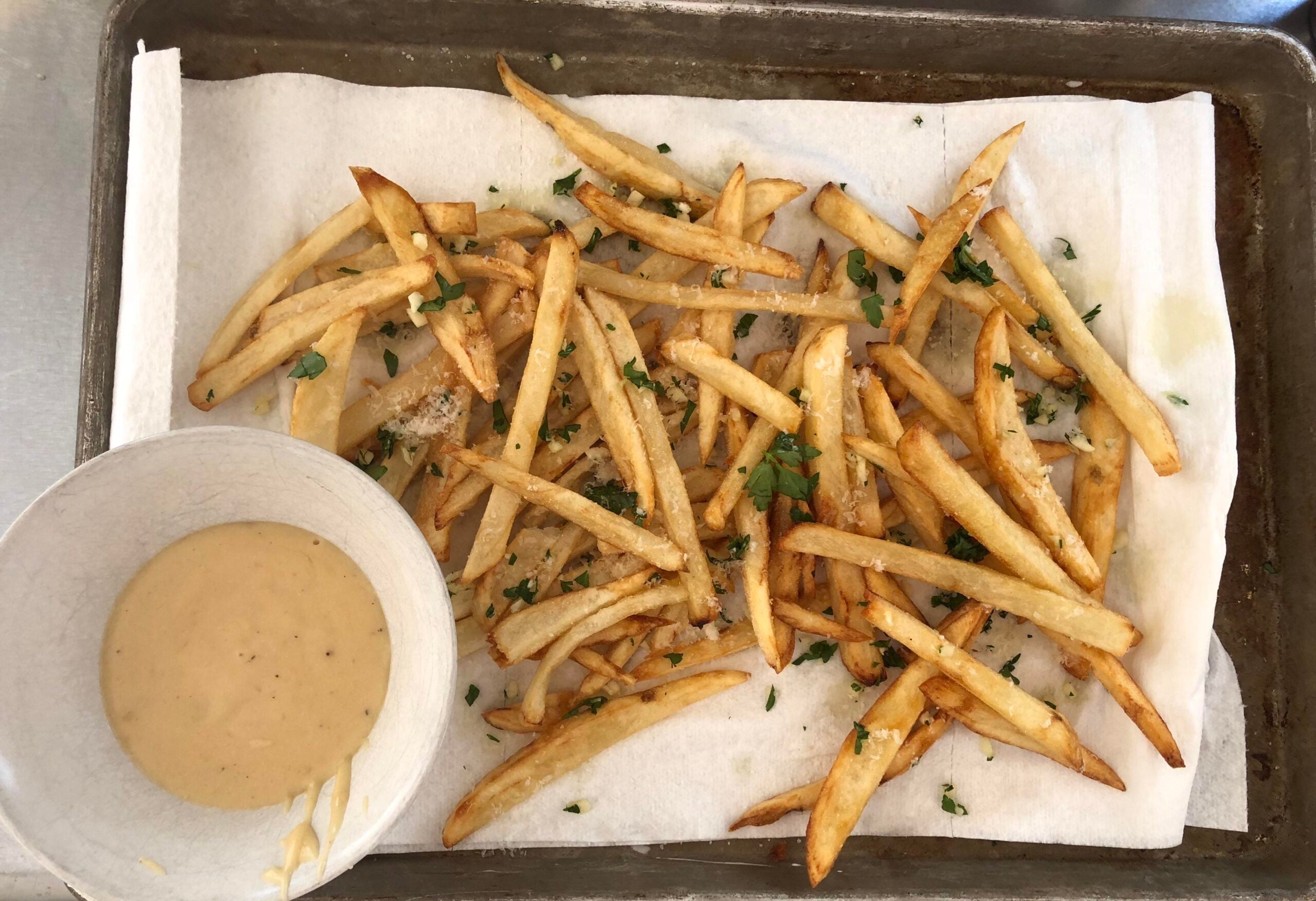 Thin French fries topped with chopped herbs spread on a paper towel-lined baking sheet, next to a small bowl of light brown dipping sauce.