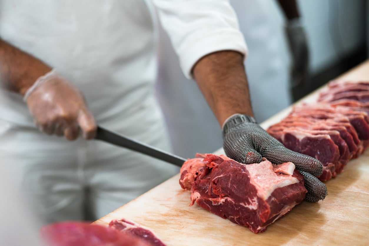 A person wearing protective gloves slices raw meat with a knife on a cutting board, with several cuts of meat lined up beside them.