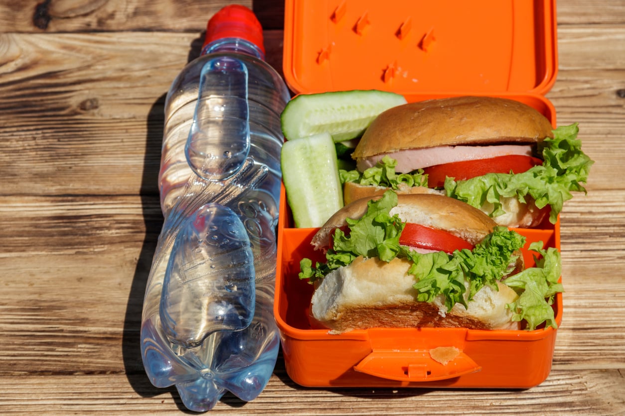 An orange lunchbox with two sandwiches, lettuce, tomato, sliced cucumber, and a plastic bottle of water on a wooden surface.