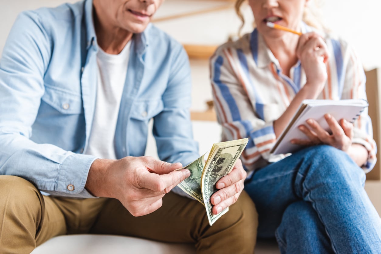 Two people sitting closely. One counts dollar bills while the other, holding a pencil and notebook, looks on thoughtfully. They appear to be discussing finances or budgeting at home.